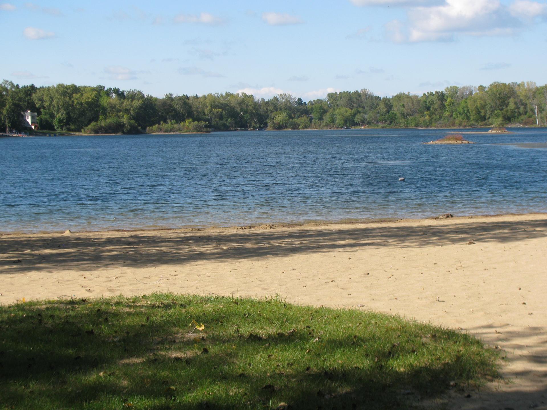 A sandy beach and grassy area next to a wide blue lake.