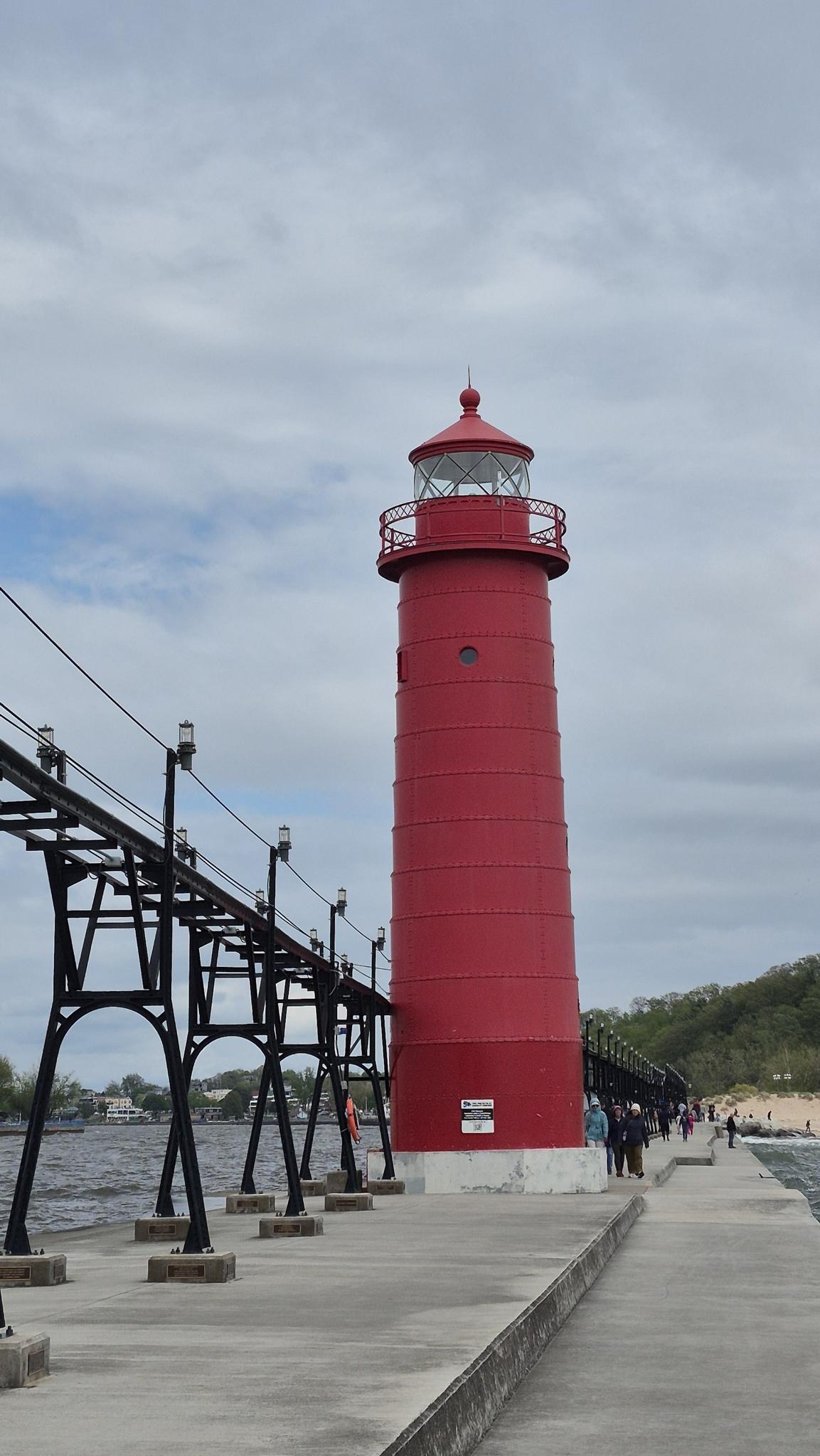 Tall red lighthouse next to a pier with distant people under a cloudy sky.