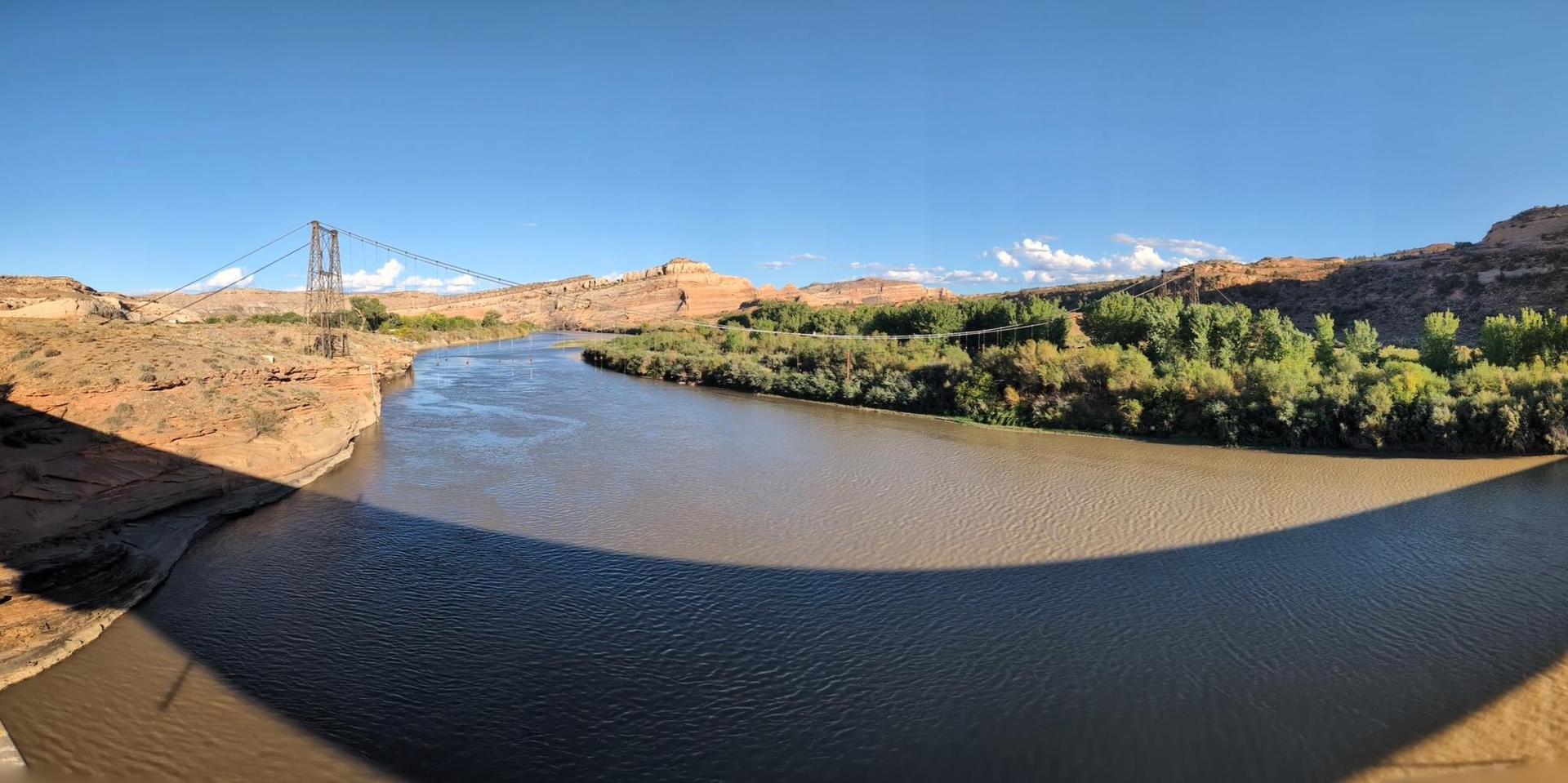 Wide river flowing through a sunny canyon with a distant suspension bridge.