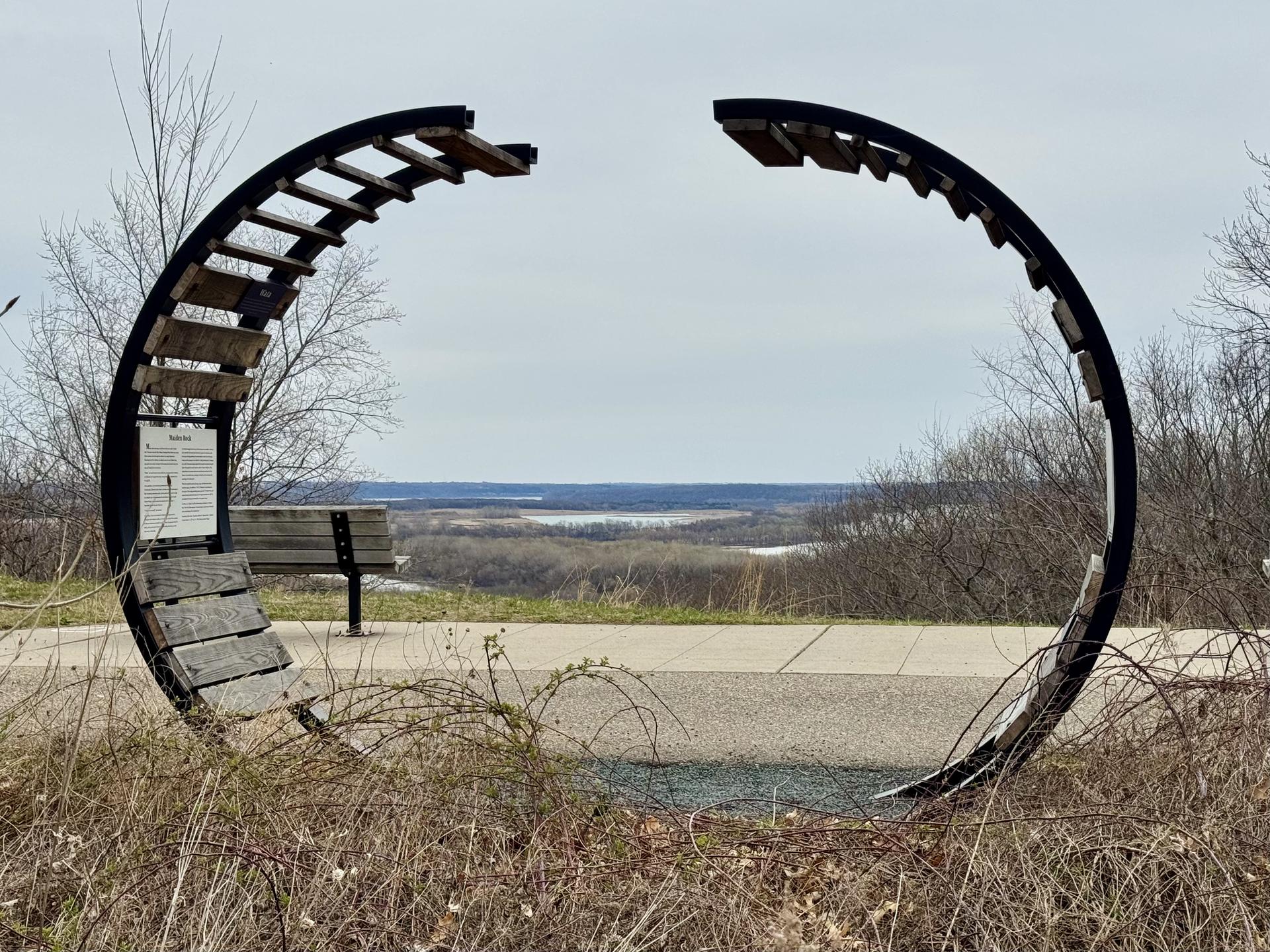 A large circular metal sculpture with wooden slats overlooks a distant valley.
