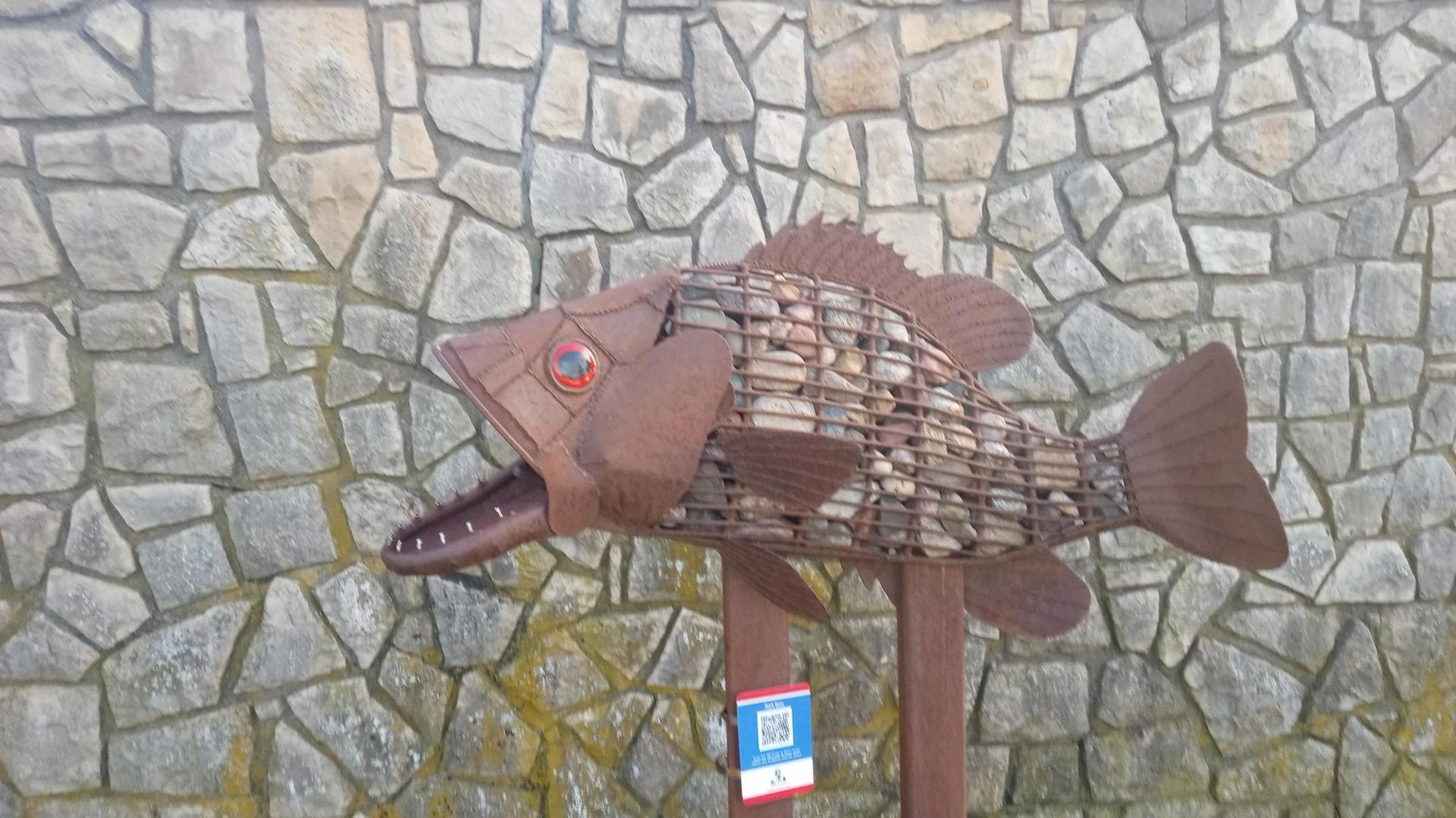 Large brown fish sculpture made of metal and stones against a stone wall.