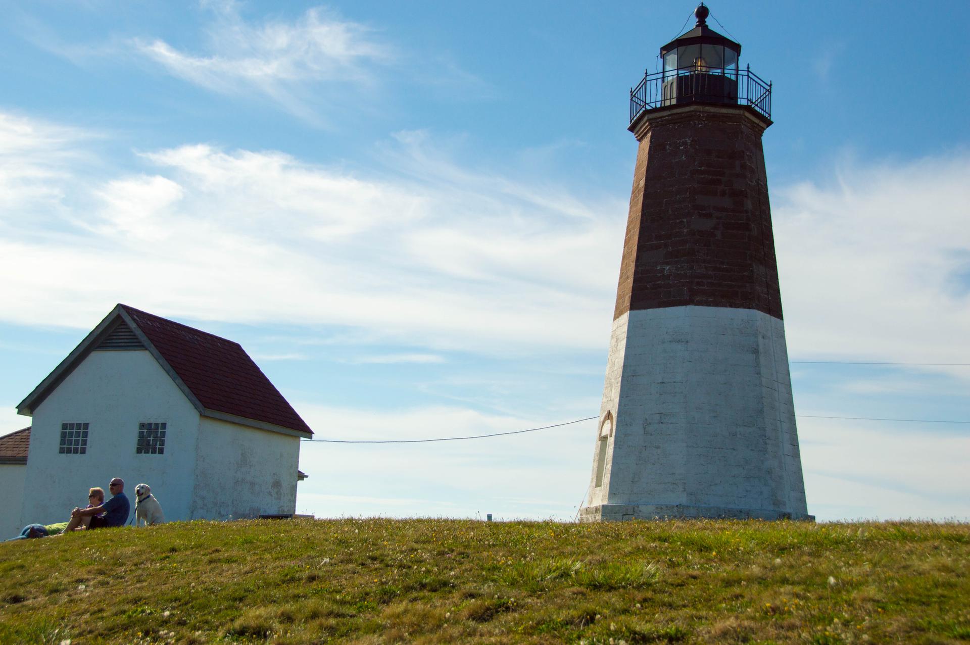 A red and white lighthouse, building, and people on a sunny grassy hill.