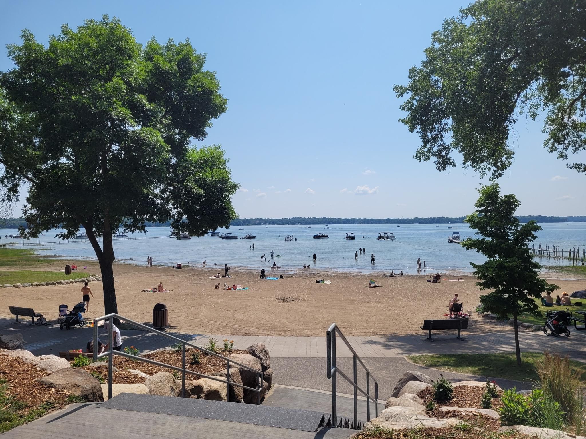 Sunny beach scene with people, boats on a lake, and green trees. - Lake Links Trail