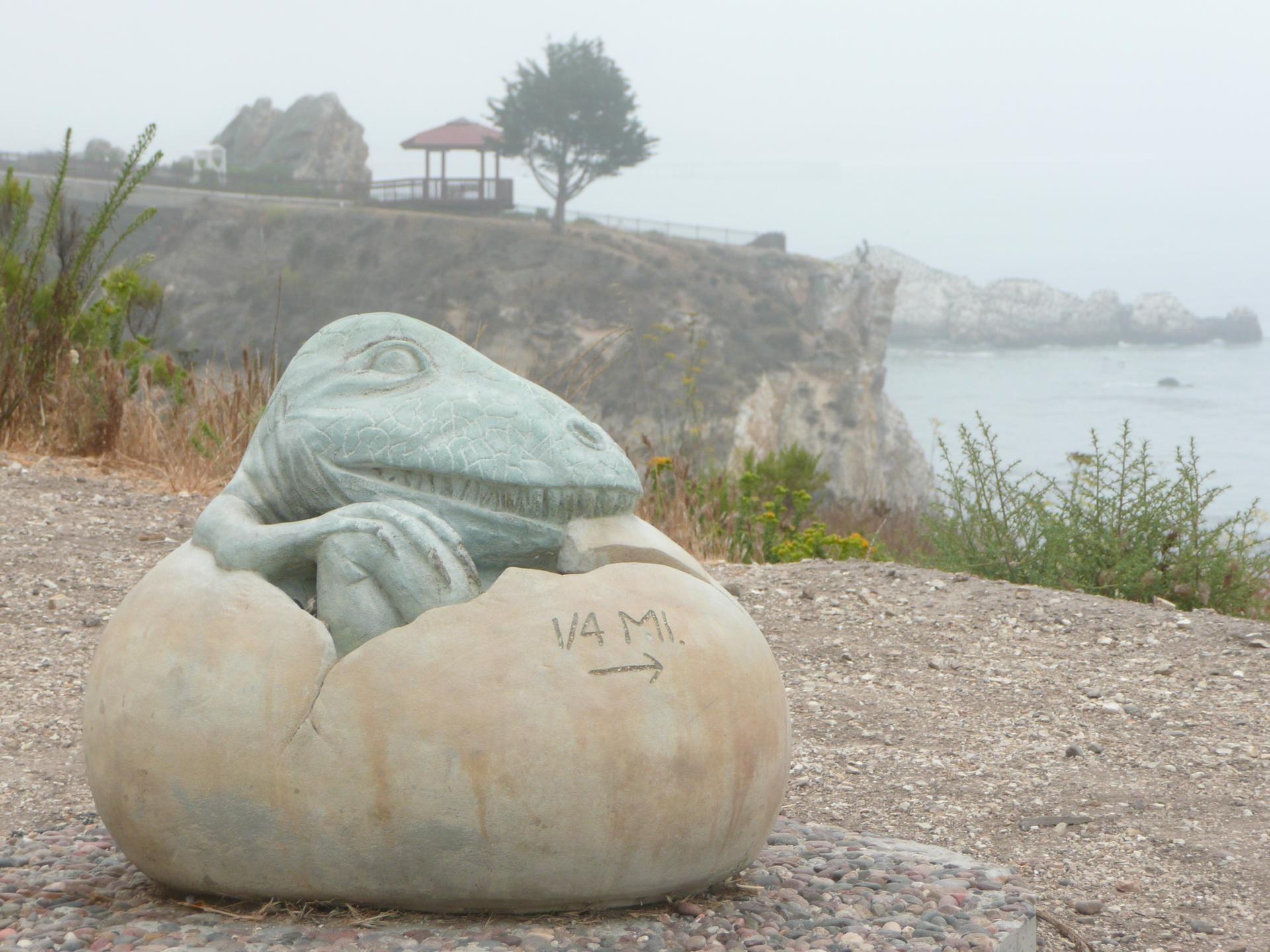 Dinosaur hatching from an egg statue on a foggy coastal cliff.