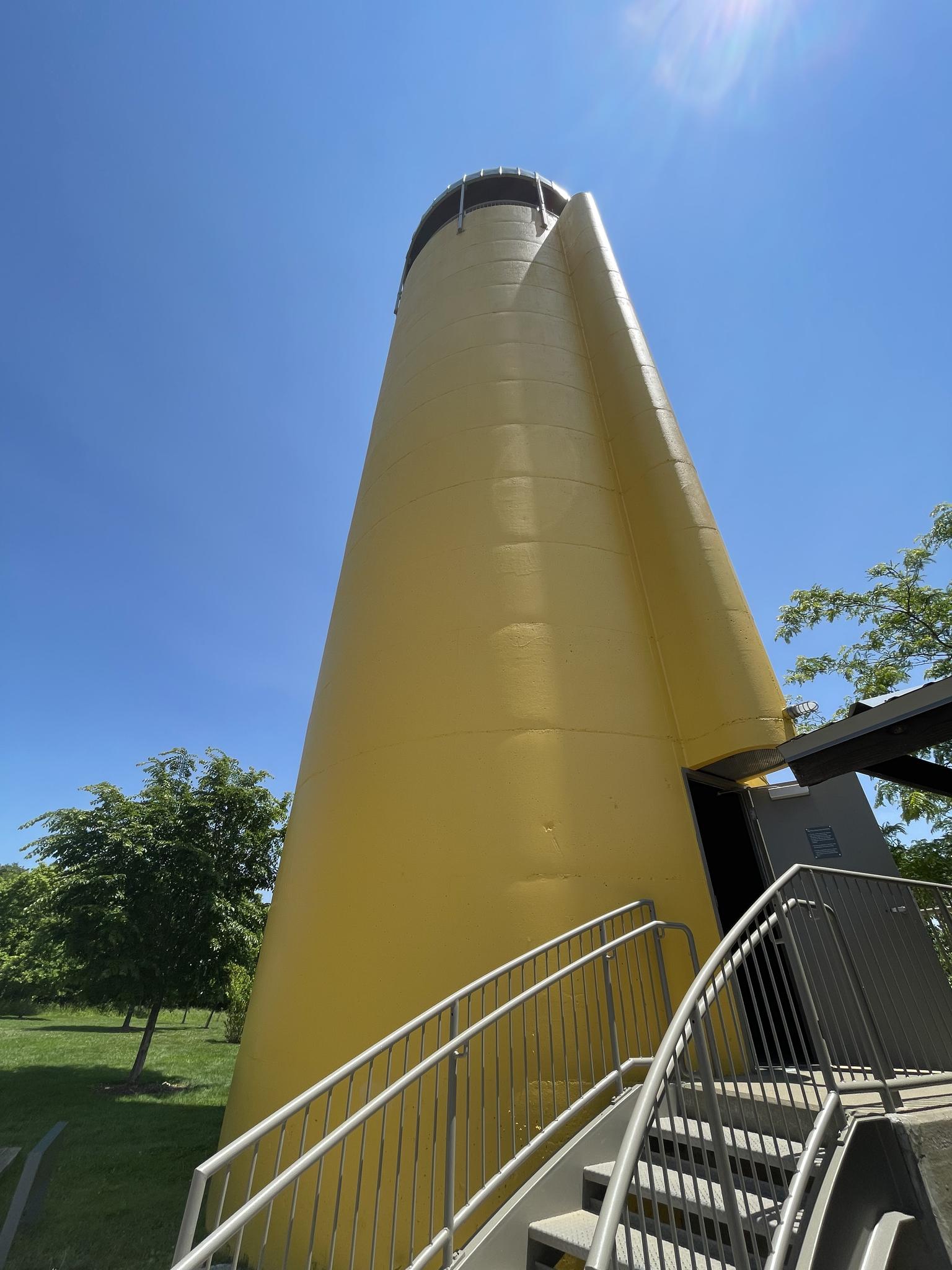 A tall, yellow, conical structure with stairs under a bright blue sky.