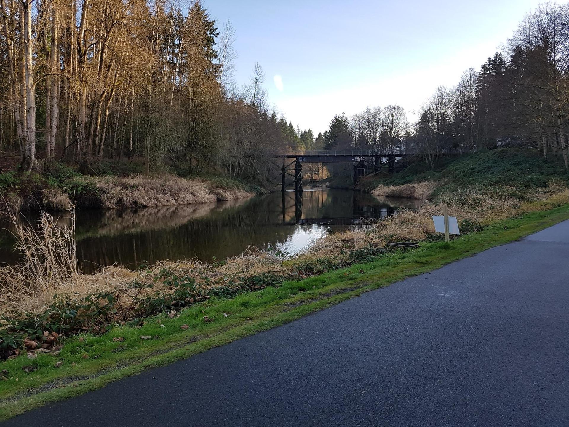 River with a dark bridge, paved path, and bare trees under a clear sky.
