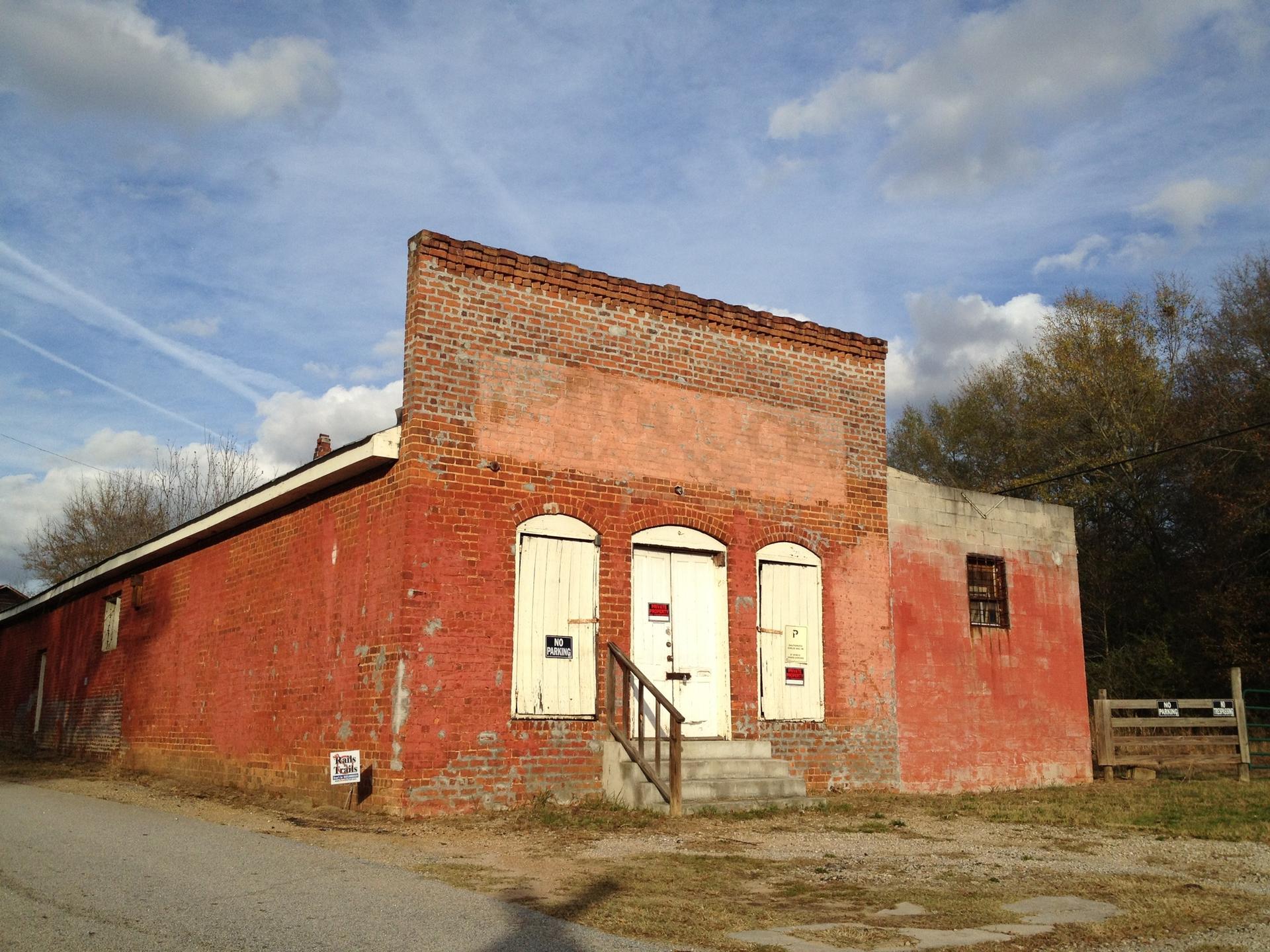 Dilapidated red brick building with boarded windows under a partly cloudy sky.