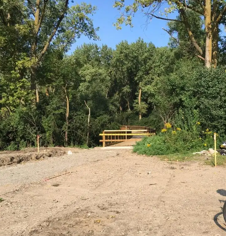 A dirt path leads to a new wooden bridge surrounded by green trees.
