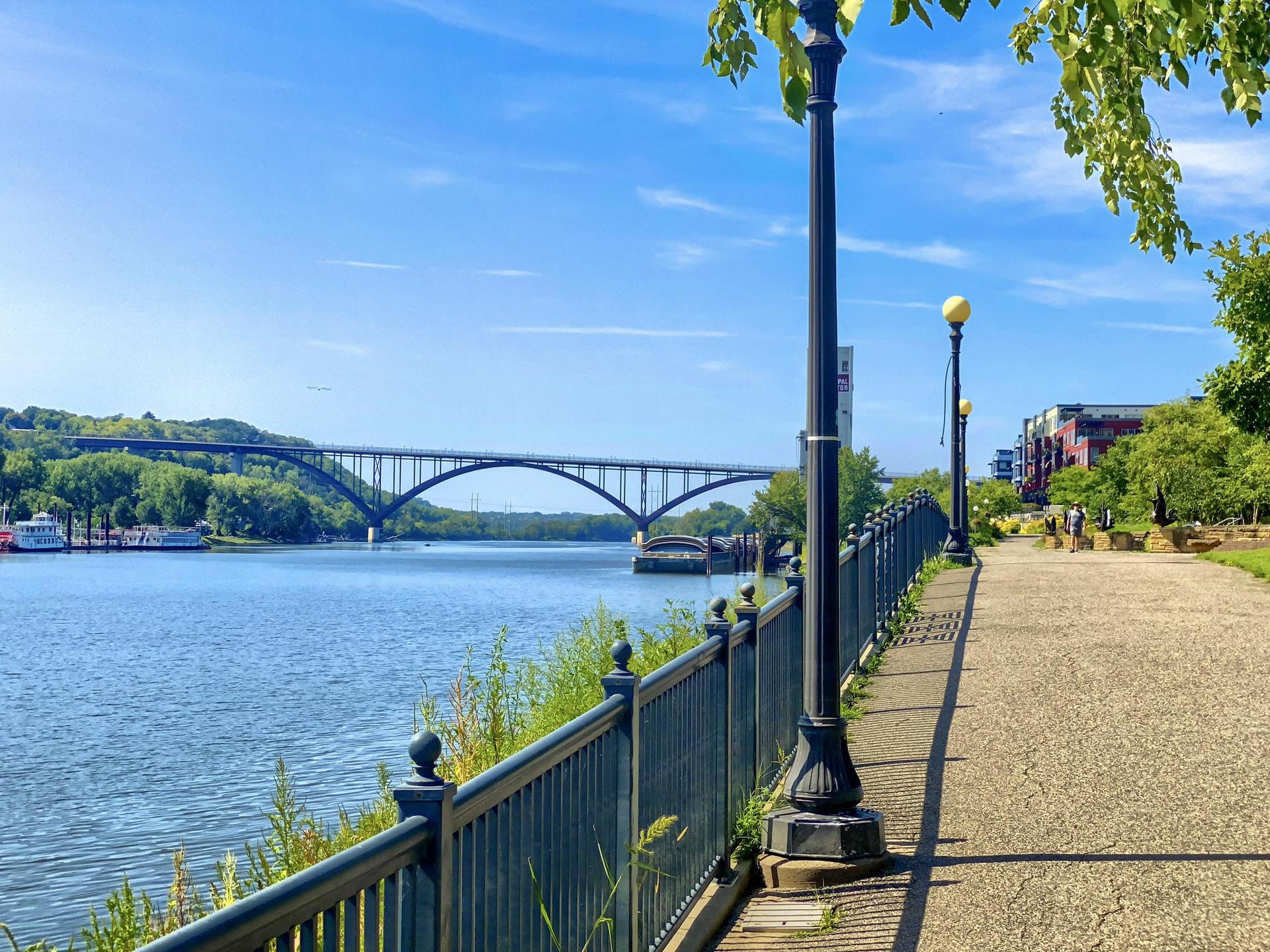 Sunny river scene with a large bridge, paved path, and green trees.