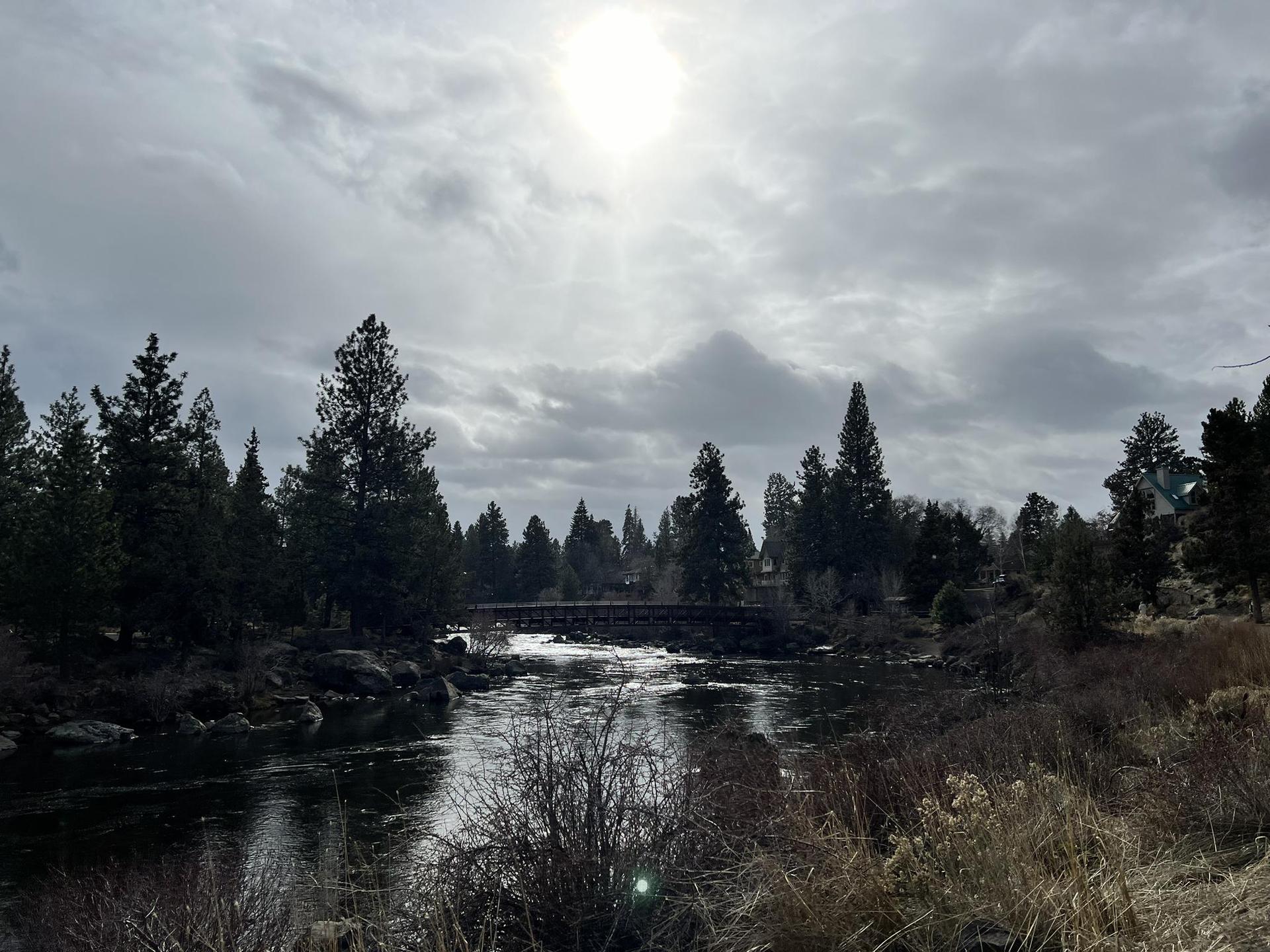 Sun breaking through clouds over a river, stone bridge, and pine trees.