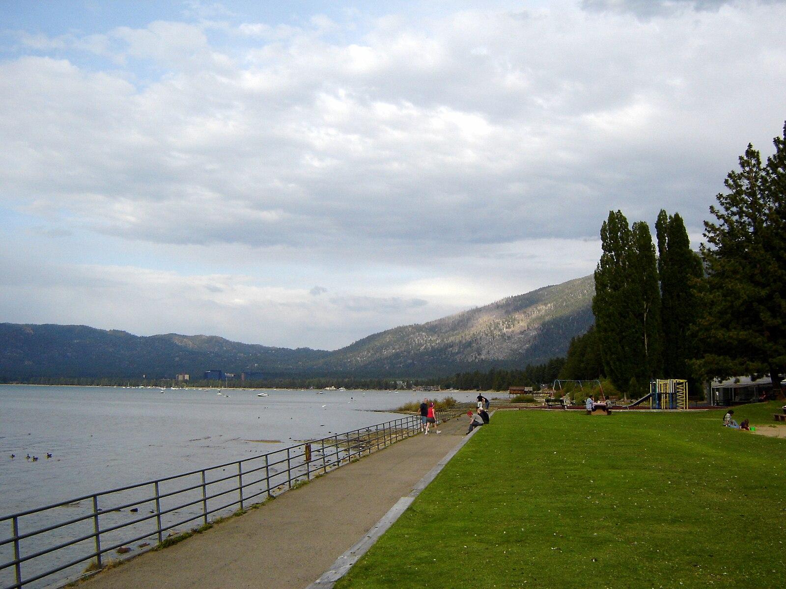 Overcast day with people walking beside a large lake and distant mountains.