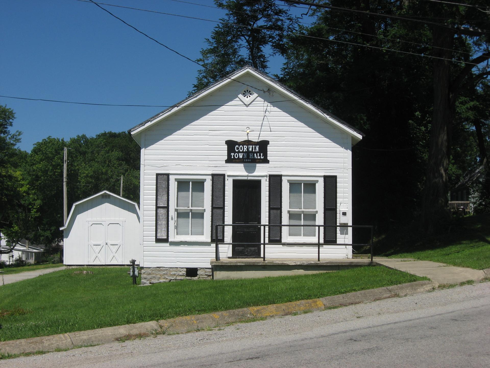 Small white town hall building with black shutters and a shed on a sunny day.