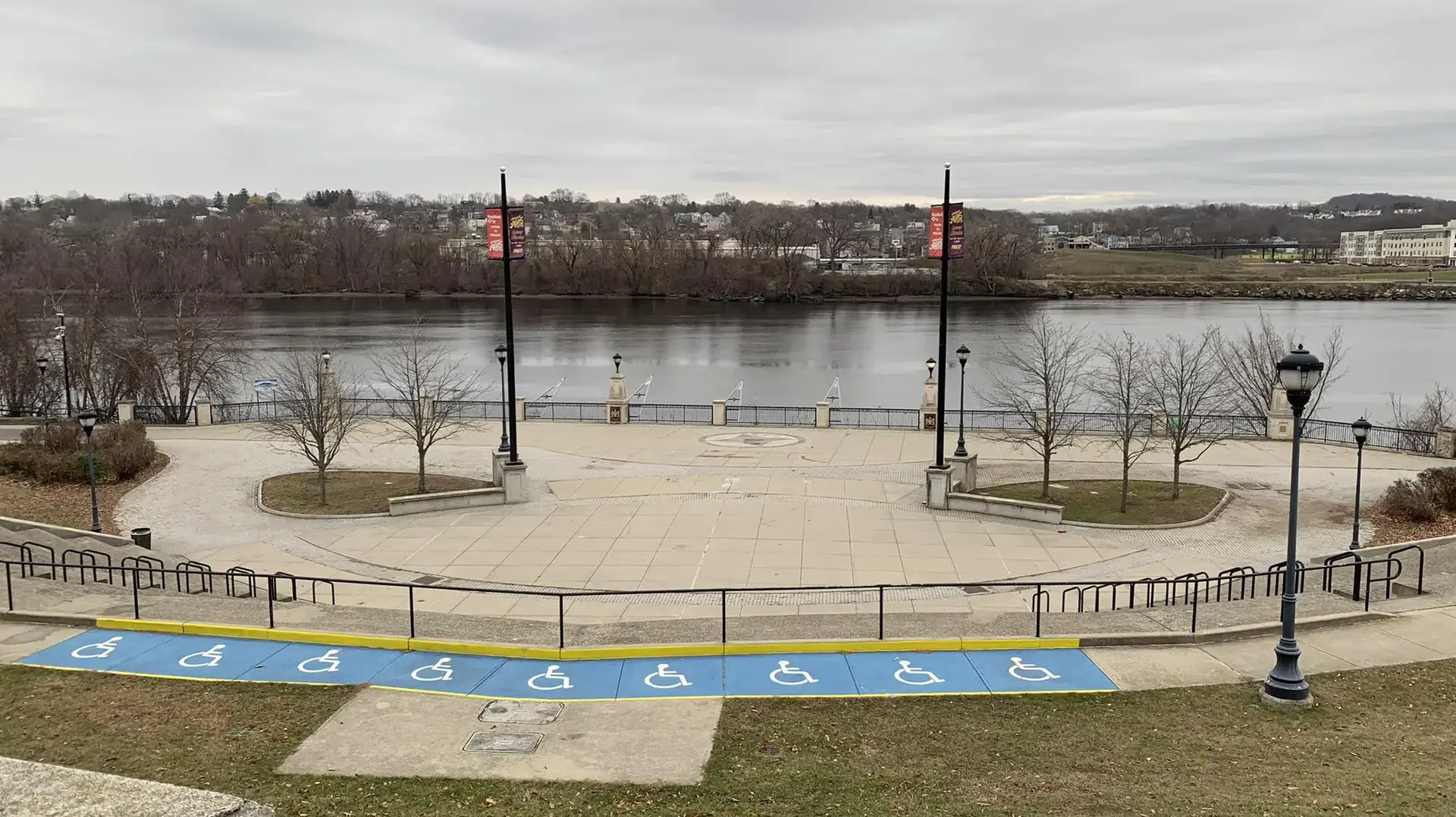 Empty waterfront plaza with a river, bare trees, and an overcast sky.