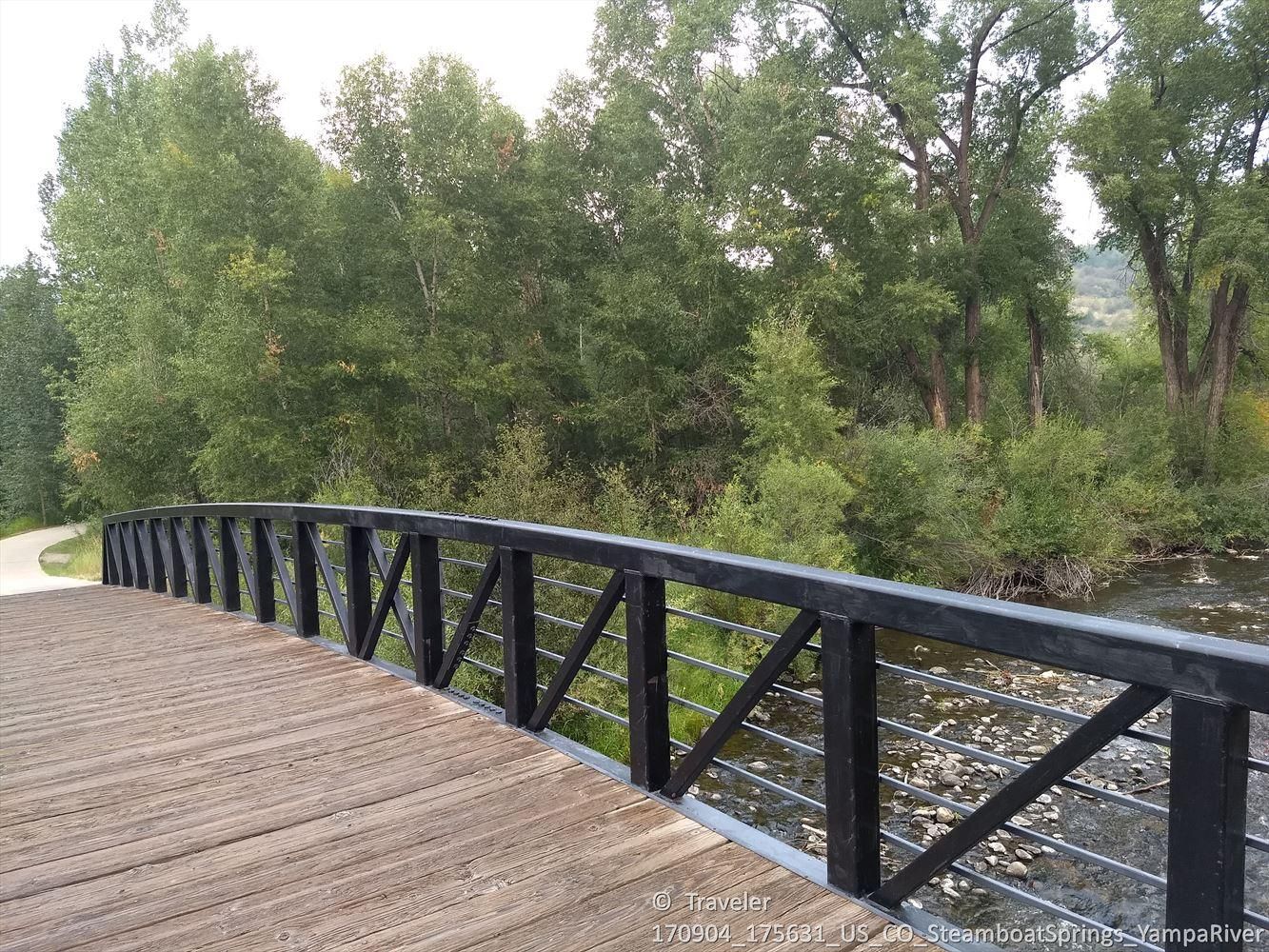 Wooden bridge with black railings crosses a river, flanked by dense green trees. - Emerald Mountain loop