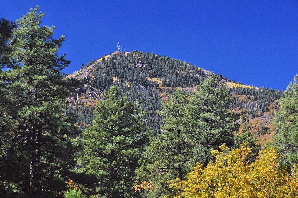 Mountain peak with communication towers, dense forest, and autumn foliage under clear sky.