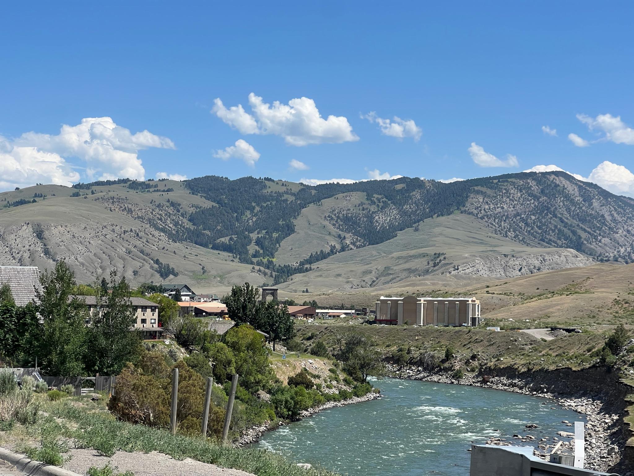 A river flows past buildings and dry mountains under a bright blue sky.