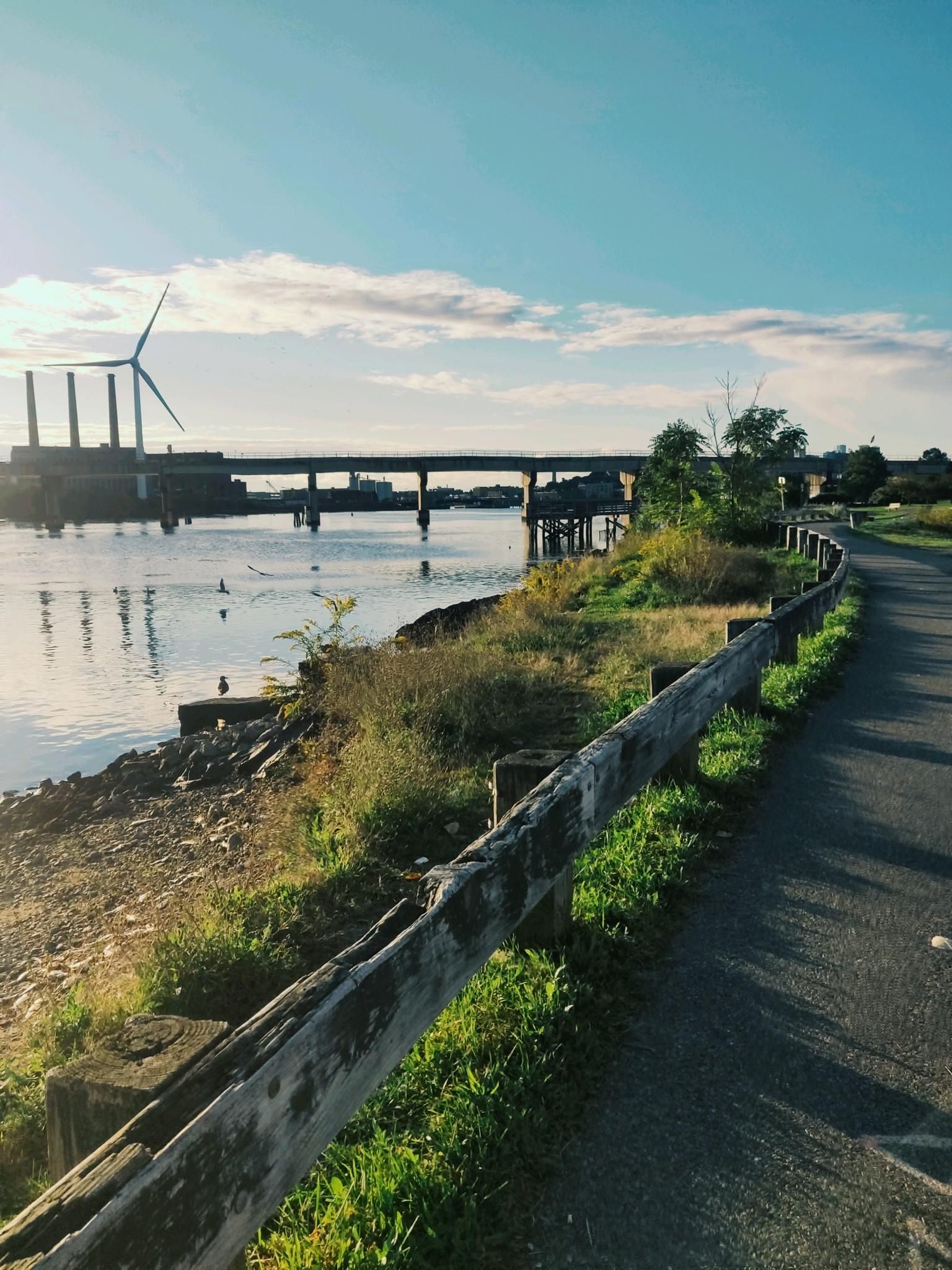 Paved path and wooden fence next to water with a distant wind turbine. - Mystic Link Trail