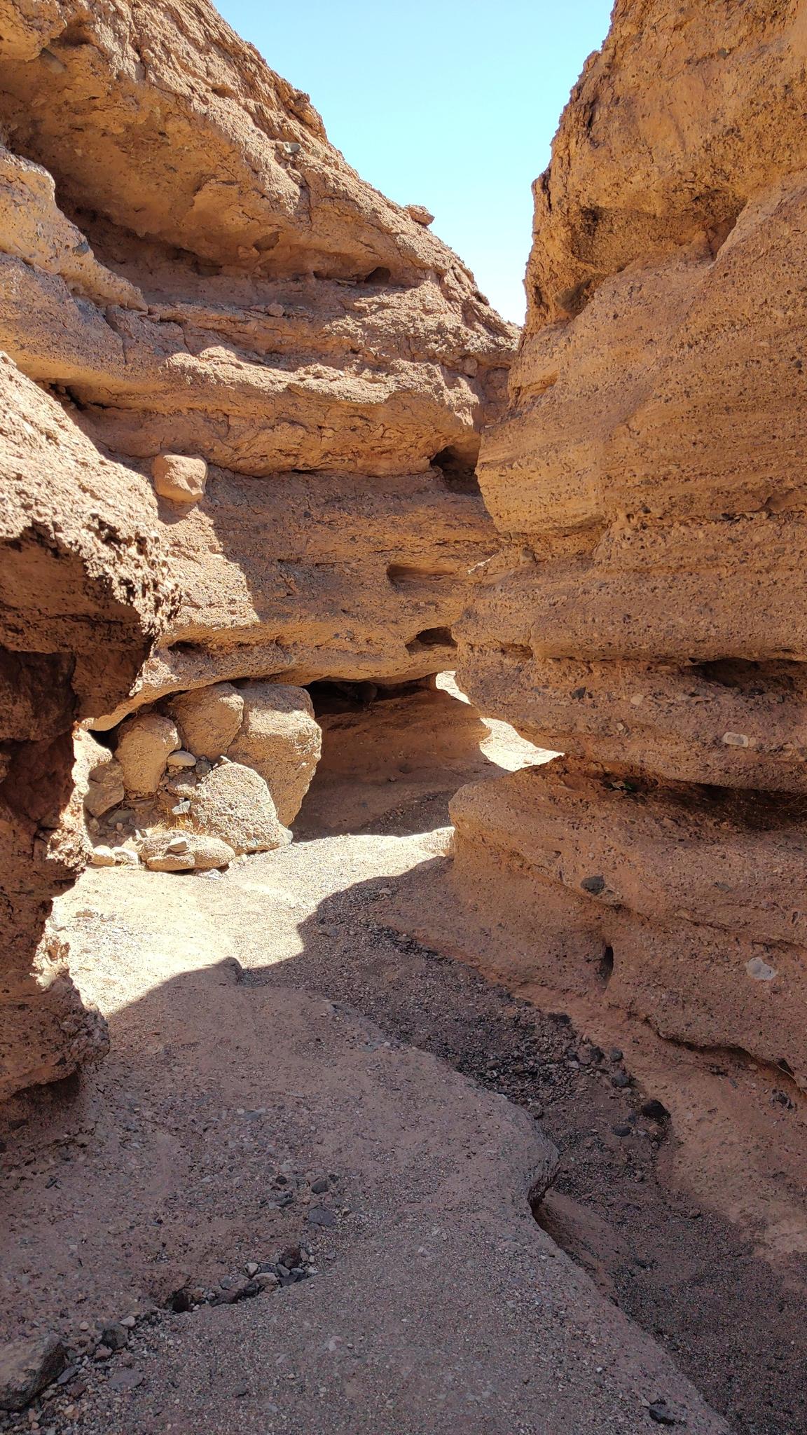 Narrow slot canyon with sunlit sandy path and towering rock walls.