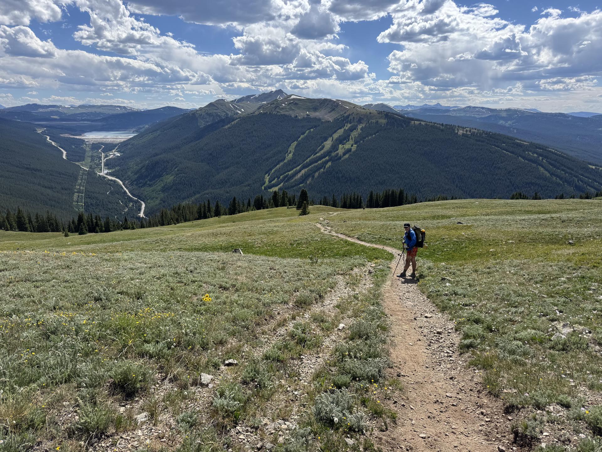 A hiker on a winding trail overlooks a vast mountain valley under clouds.
