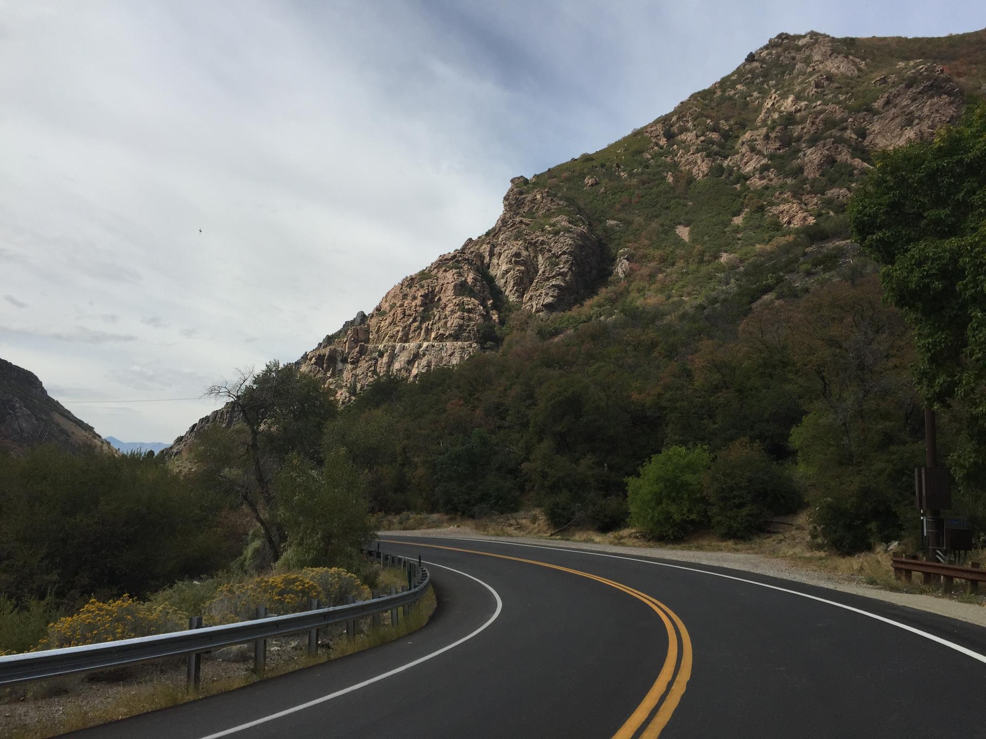 Winding road curves past a rocky, vegetated mountain under a cloudy sky.