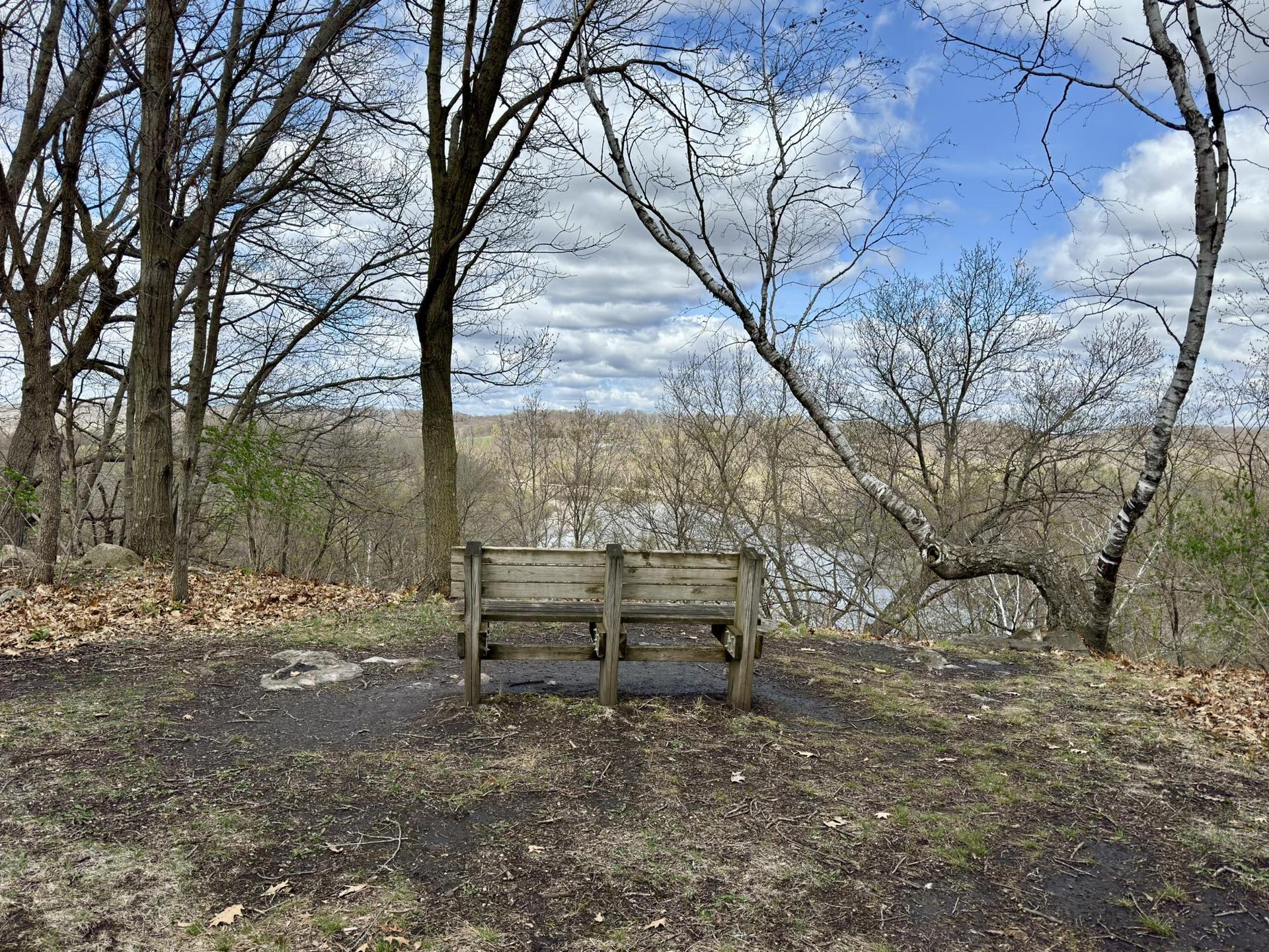 A wooden bench overlooks a distant river and cloudy sky through bare trees.