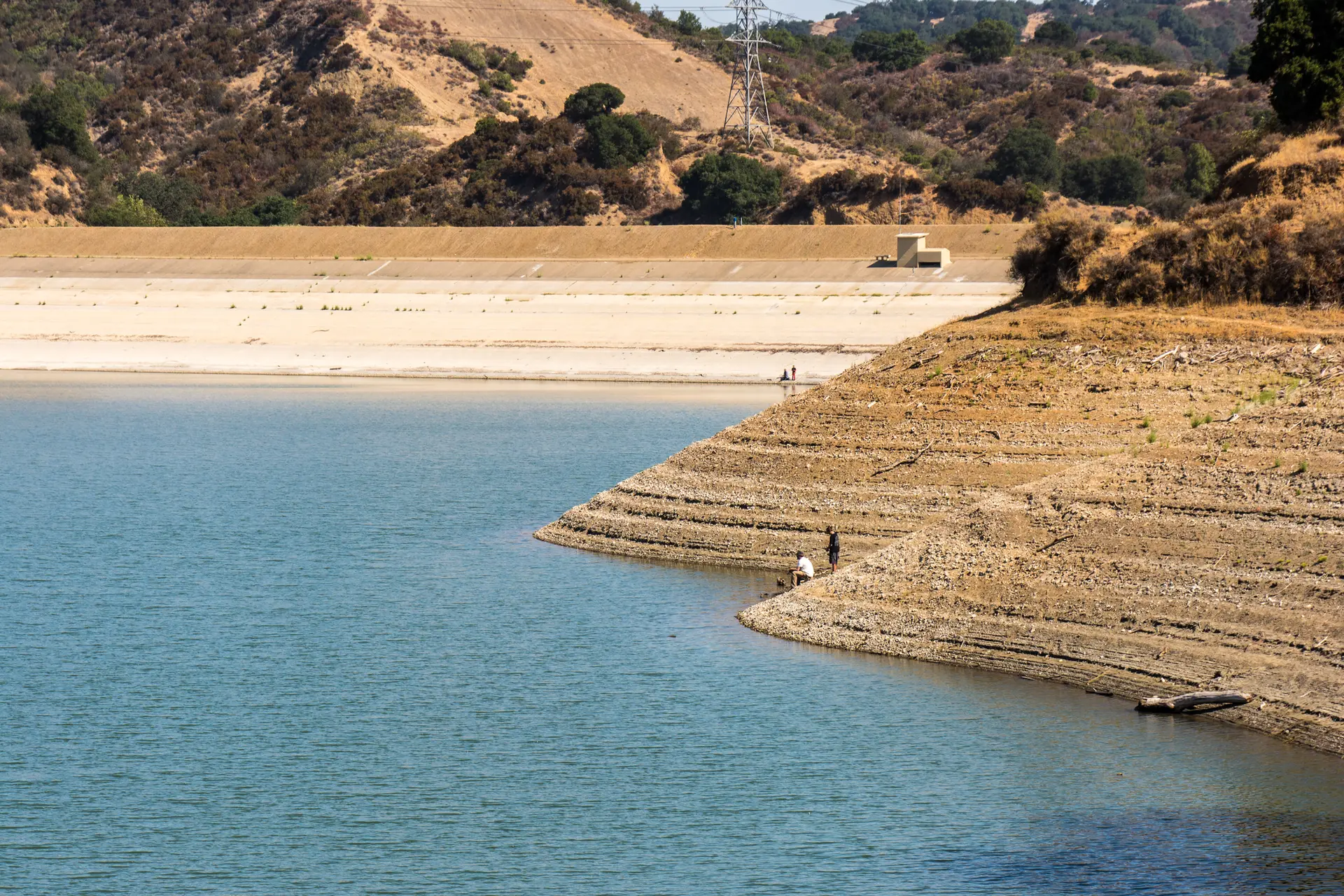 Blue water, dry terraced shoreline, distant hills, and two people fishing.