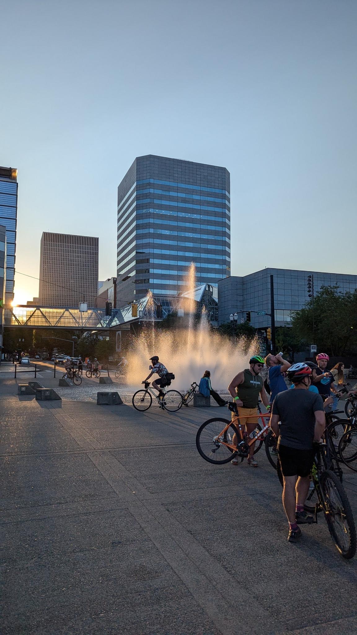 People gather with bicycles around a fountain in a city square at sunset.