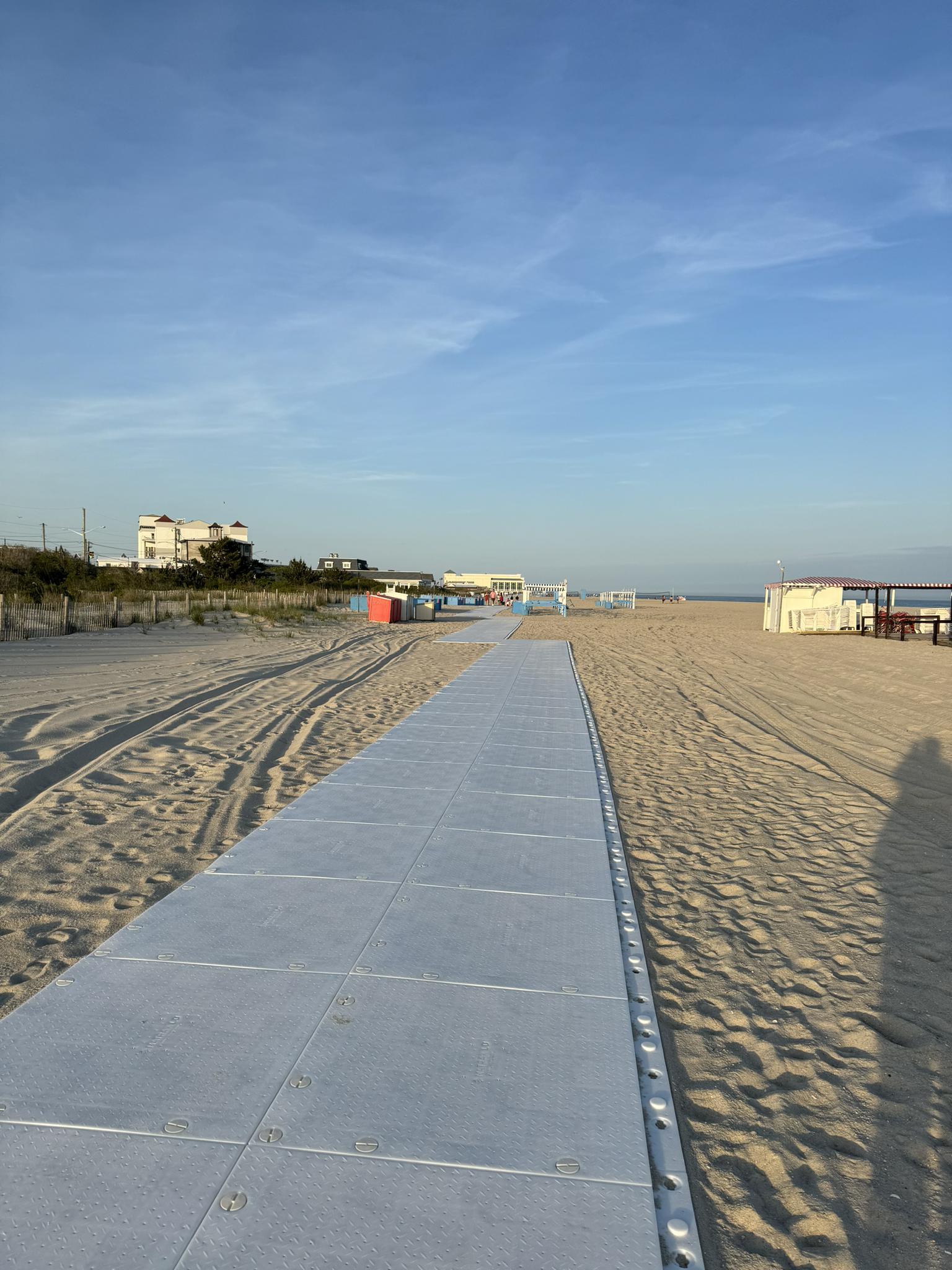 Beach access mat on sand leading to distant buildings under a blue sky.