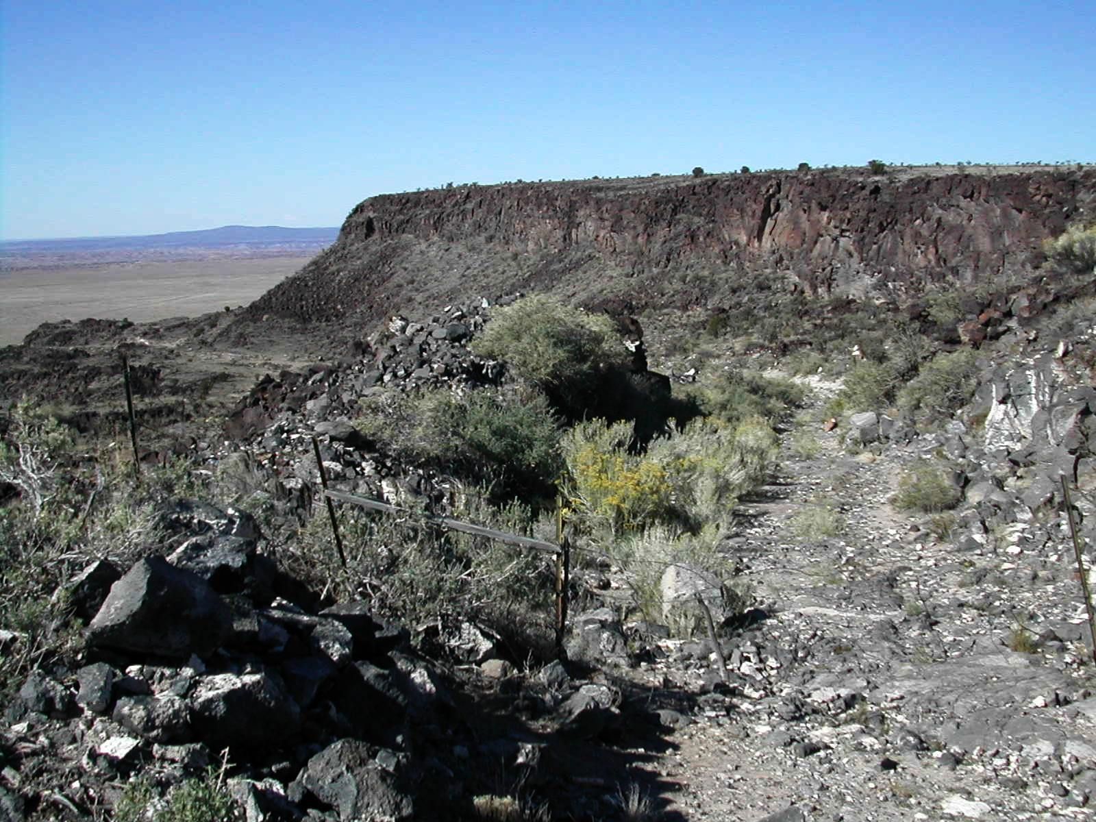 Arid, rocky landscape with a dirt path leading towards a distant mesa. - El Camino Real and Caja del Rio Loop