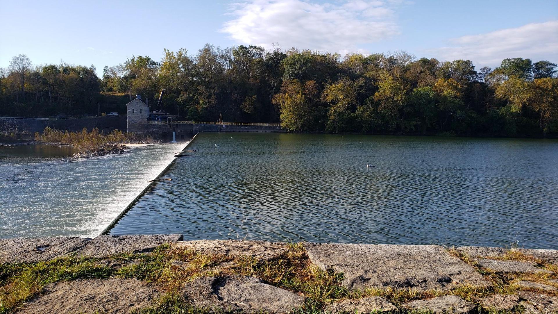 River with a low dam, small building, and autumn trees under a bright sky.