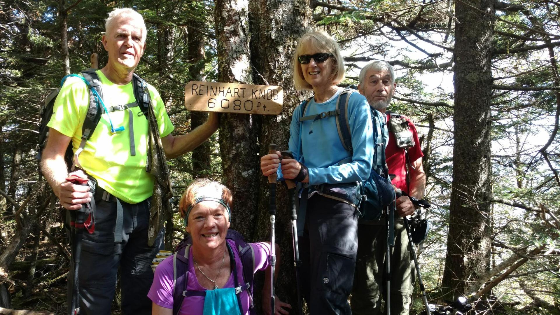 Four smiling hikers pose in a sunny forest next to a wooden sign.