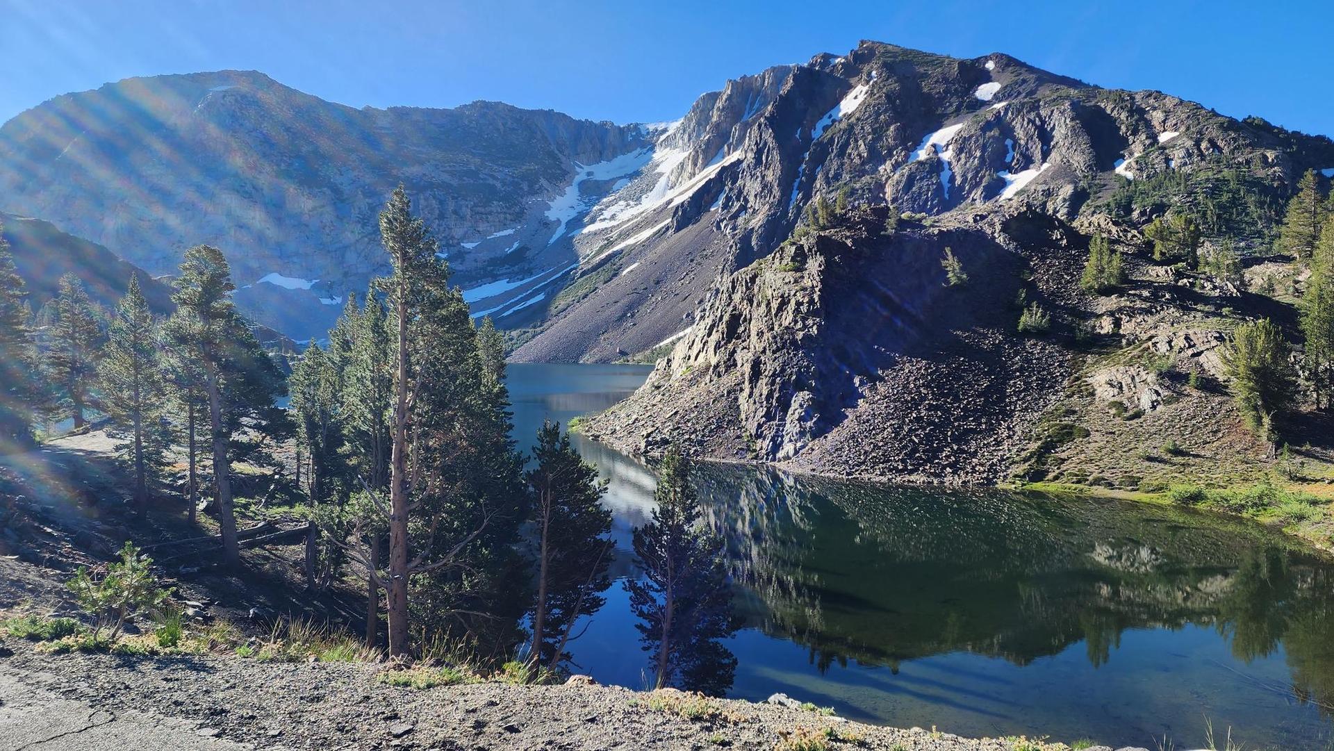 Sunny mountain lake reflecting trees, surrounded by rocky, snow-dusted peaks.