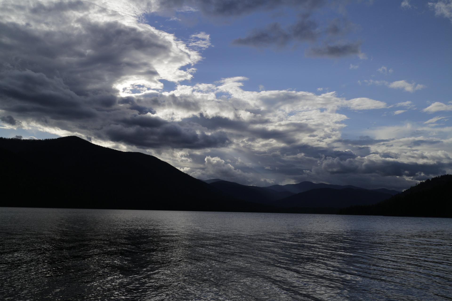 Dark mountains and a lake under a dramatic, cloudy sky with bright patches.