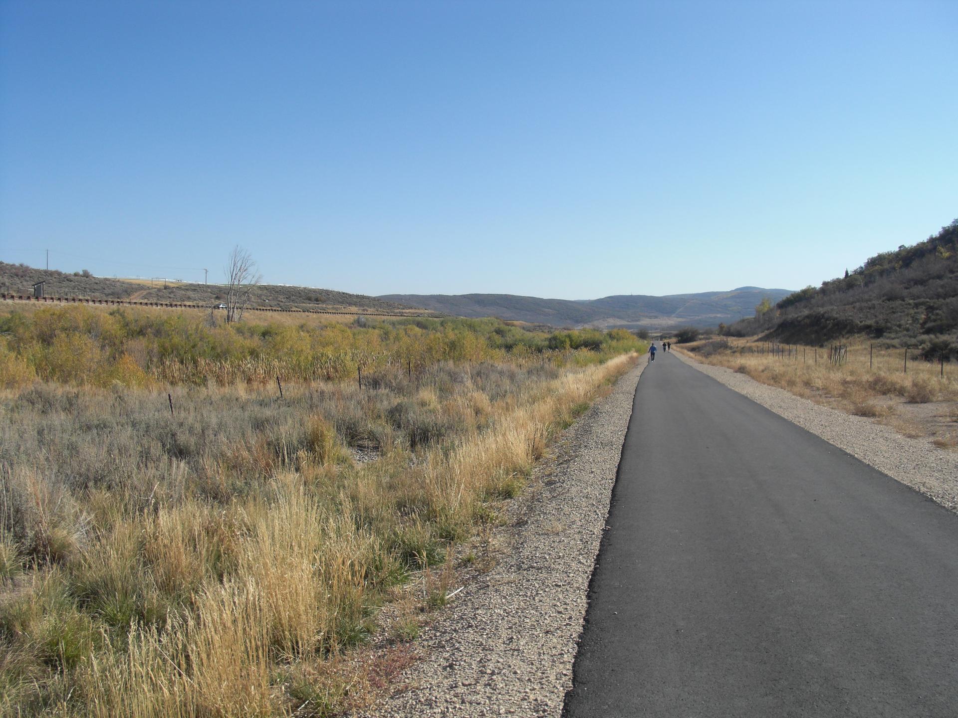Paved path through dry grass and hills, with people walking in the distance.