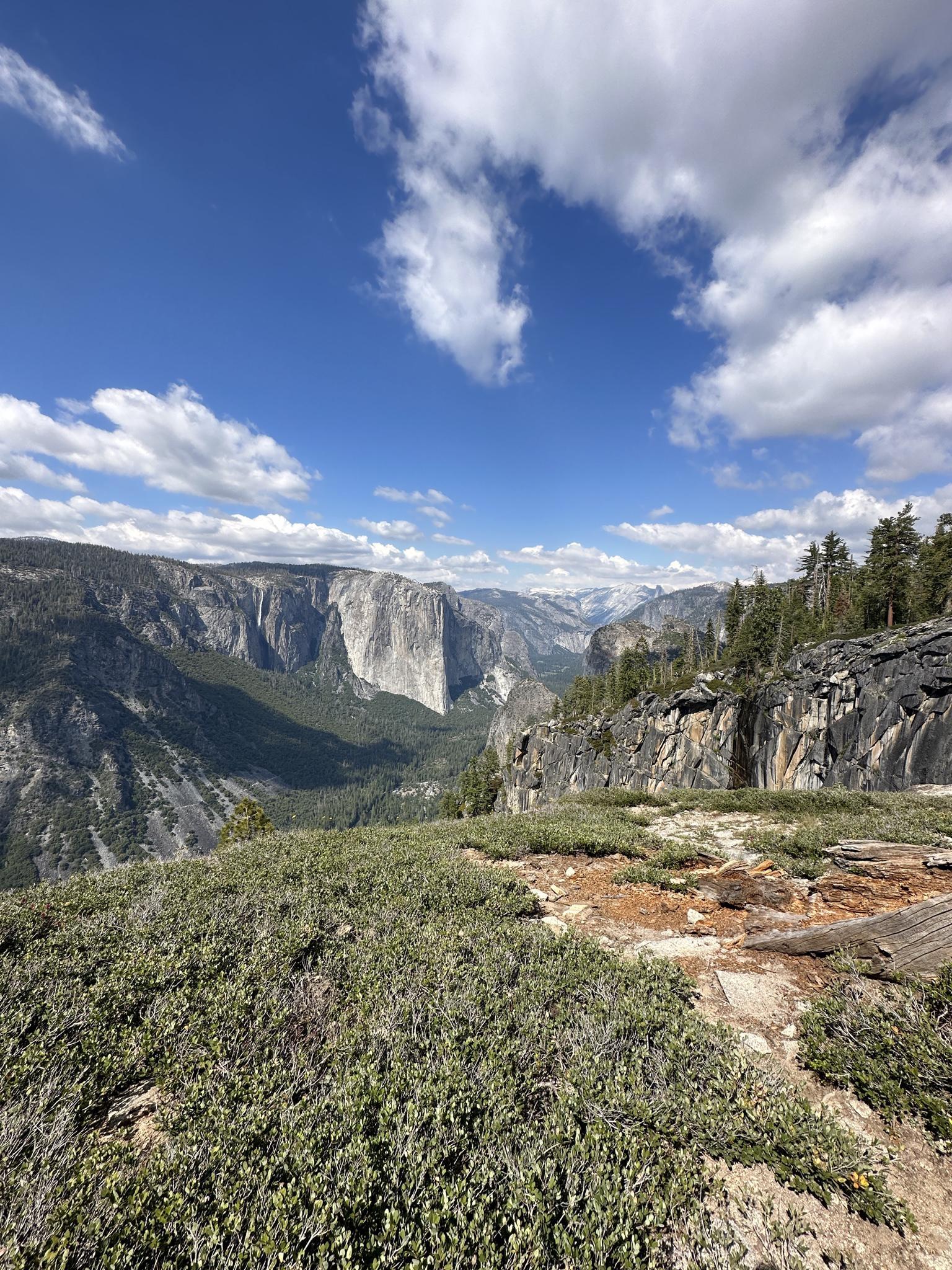 Mountain valley with a prominent rock formation, green bushes, and bright blue sky.