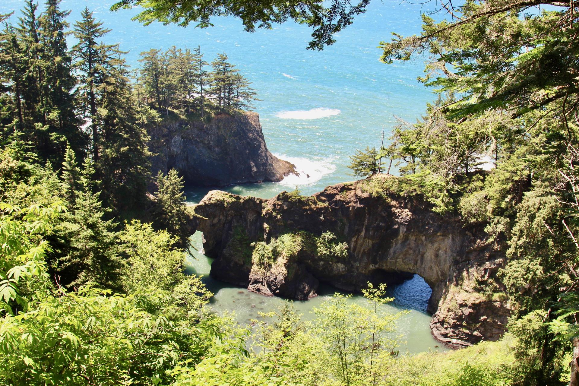Sunny view of a natural rock arch connecting forested cliffs over a calm cove.