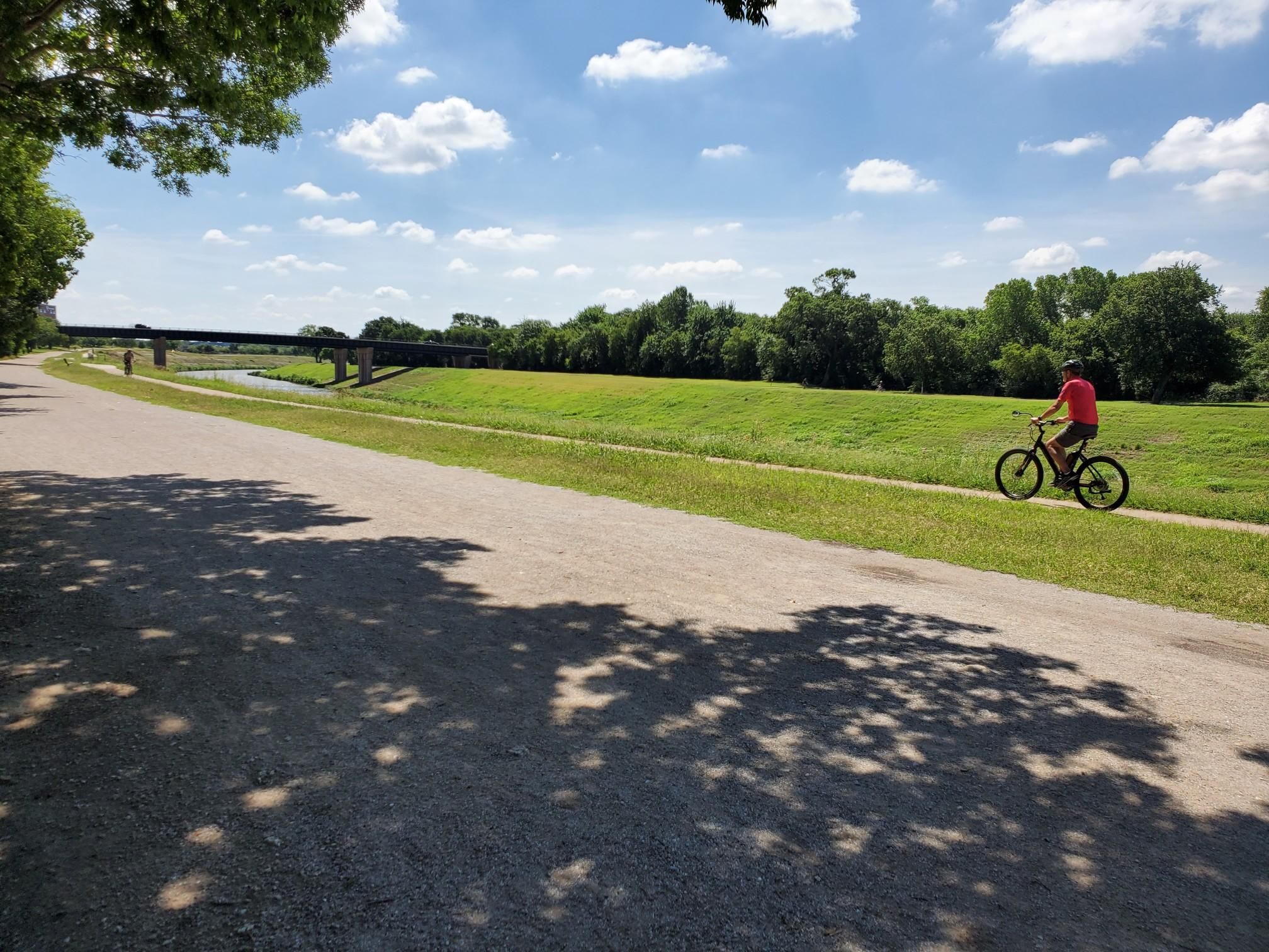 A person cycles on a sunny dirt path next to a grassy field. - Trinity Trails