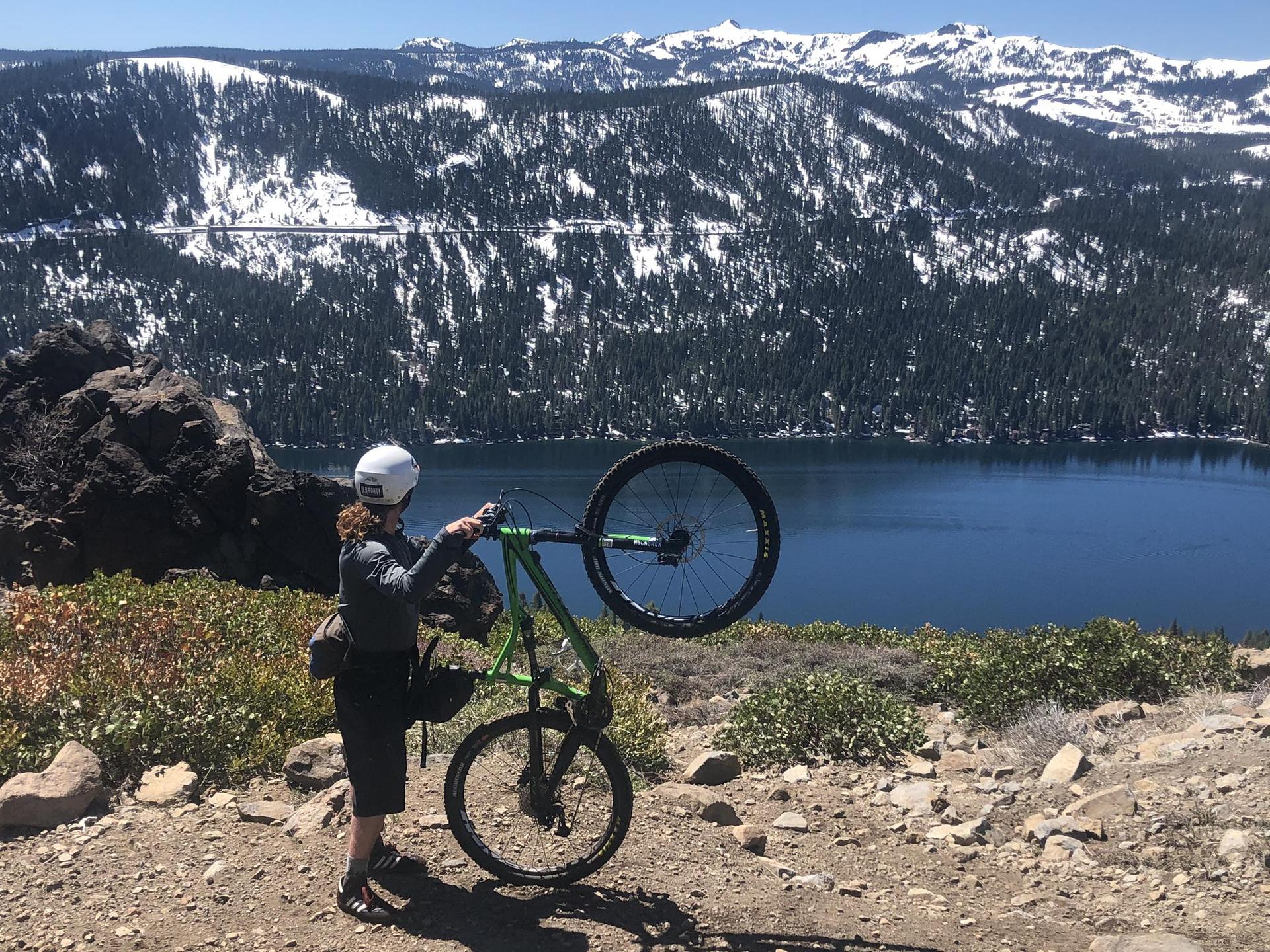 Person in helmet holding a green mountain bike overlooking a lake and snowy mountains.