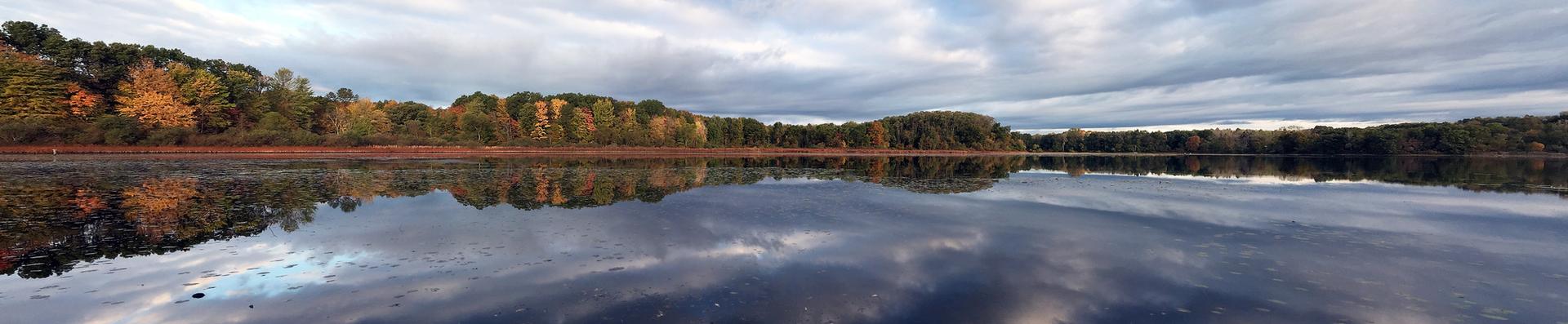 Panoramic view of a calm lake reflecting cloudy sky and autumn trees.
