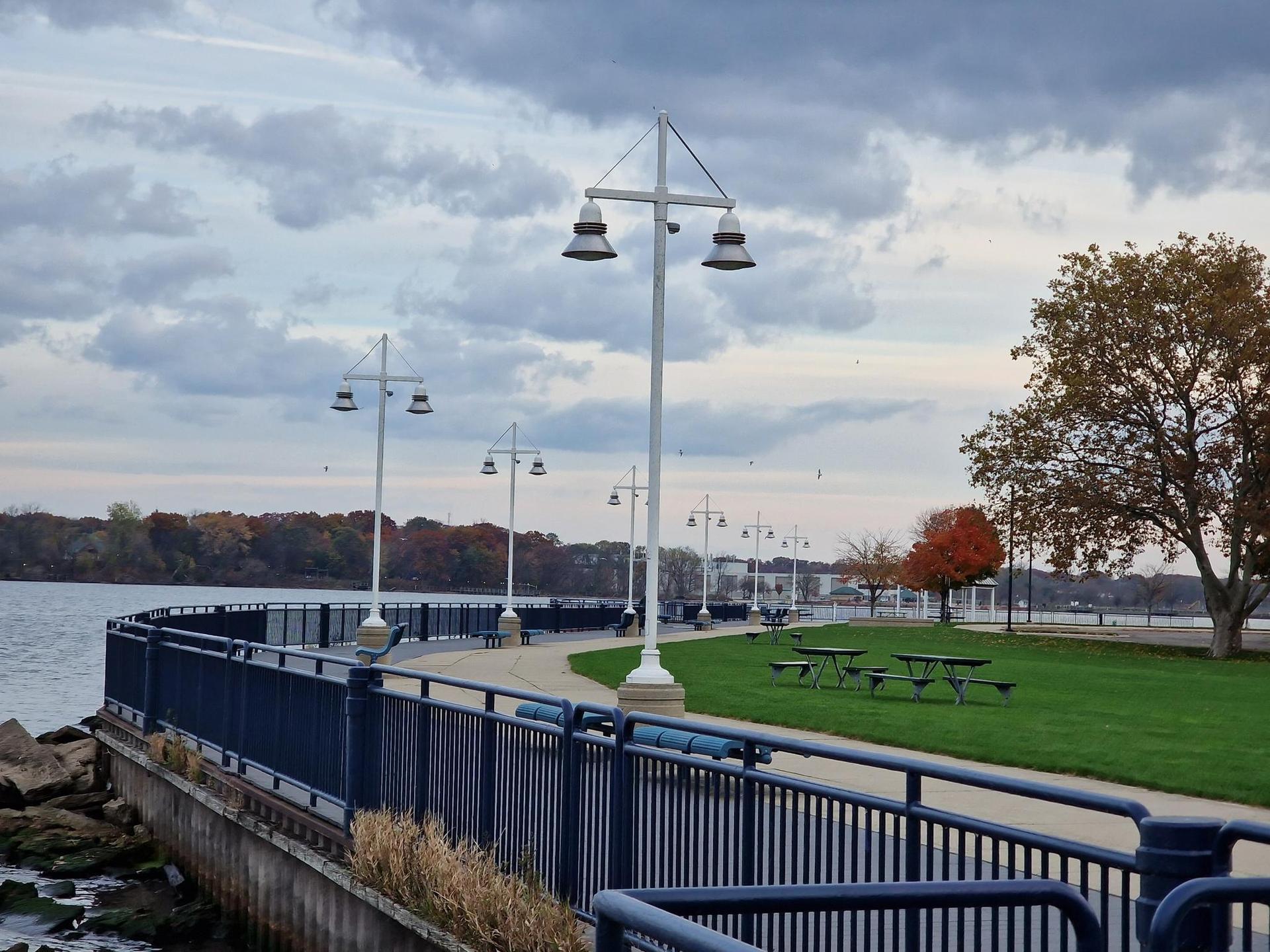 Waterfront promenade with lampposts, dark railing, and grassy park under a cloudy sky.
