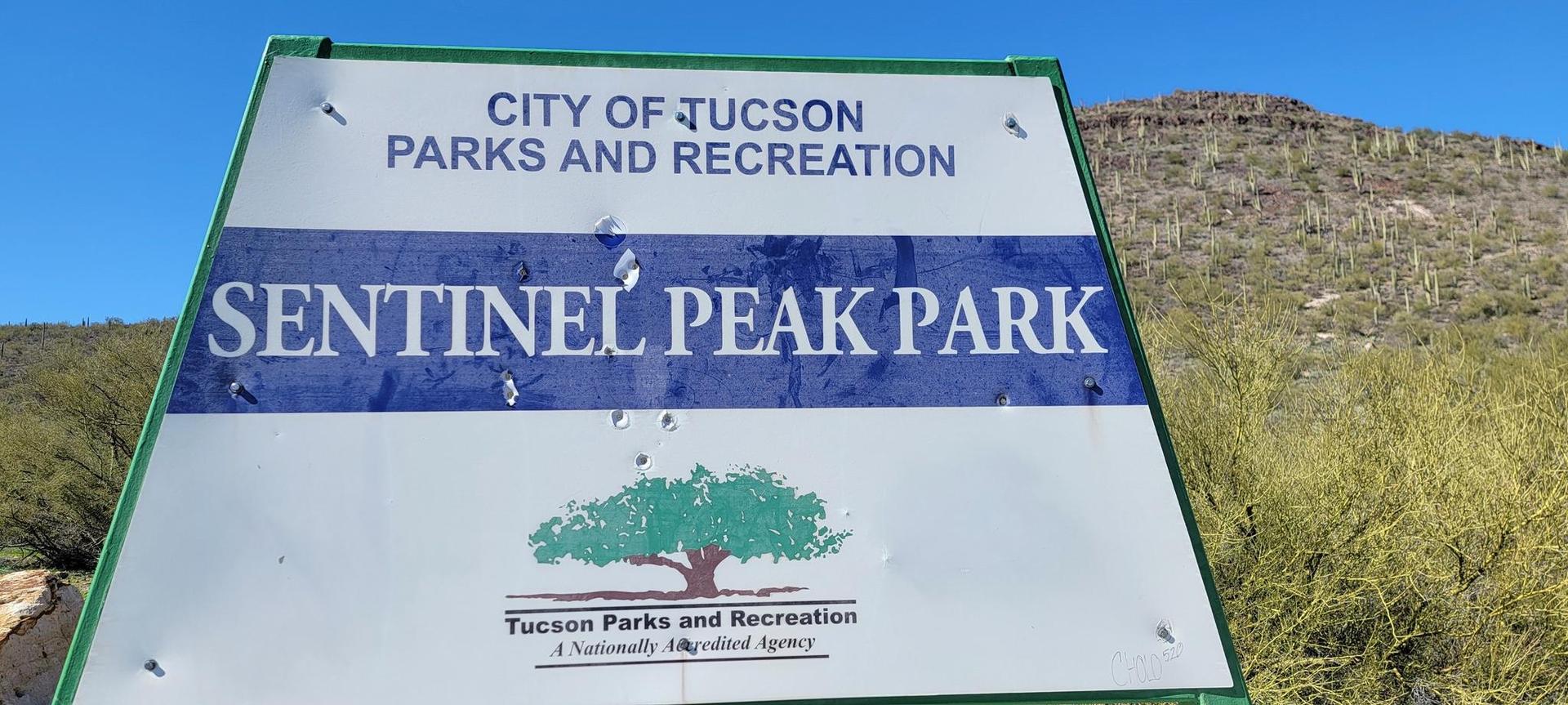 White and green park sign in bright desert sun, rocky hills behind.