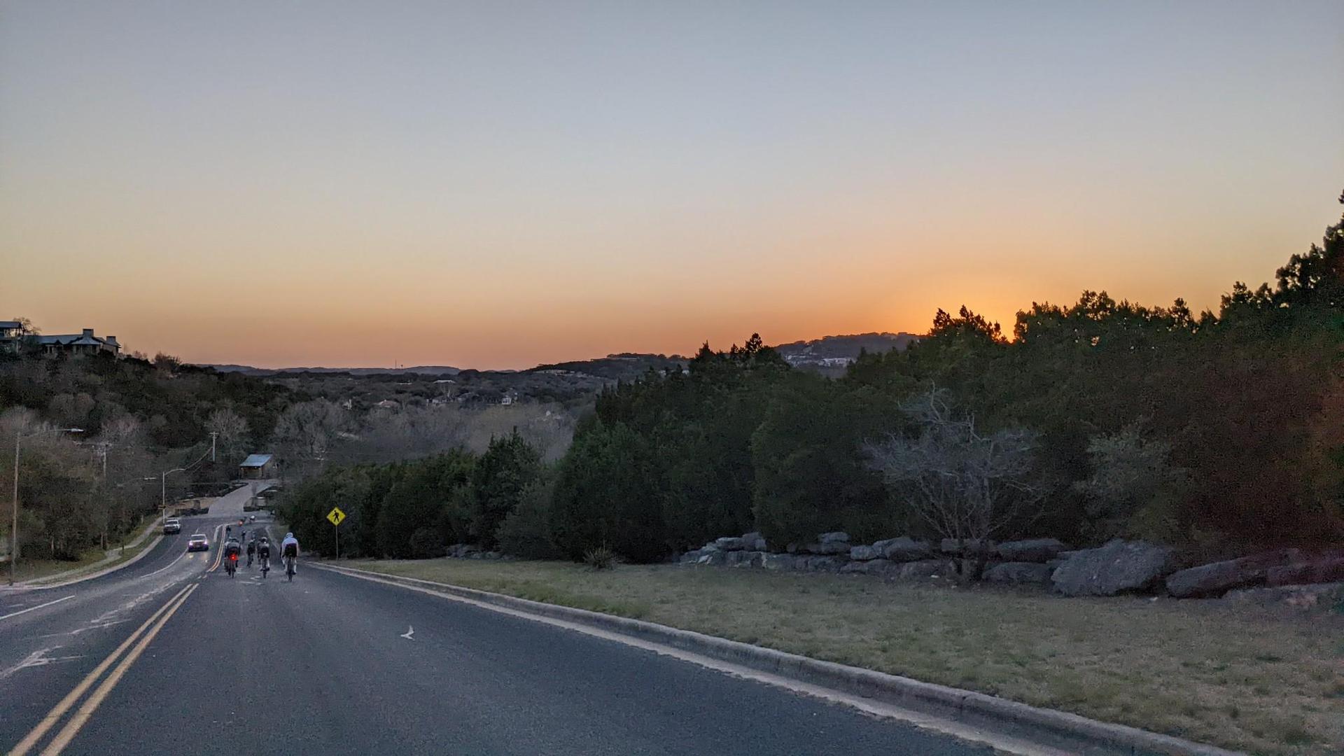 Cyclists ride on a road at dusk with a warm sunset glow.