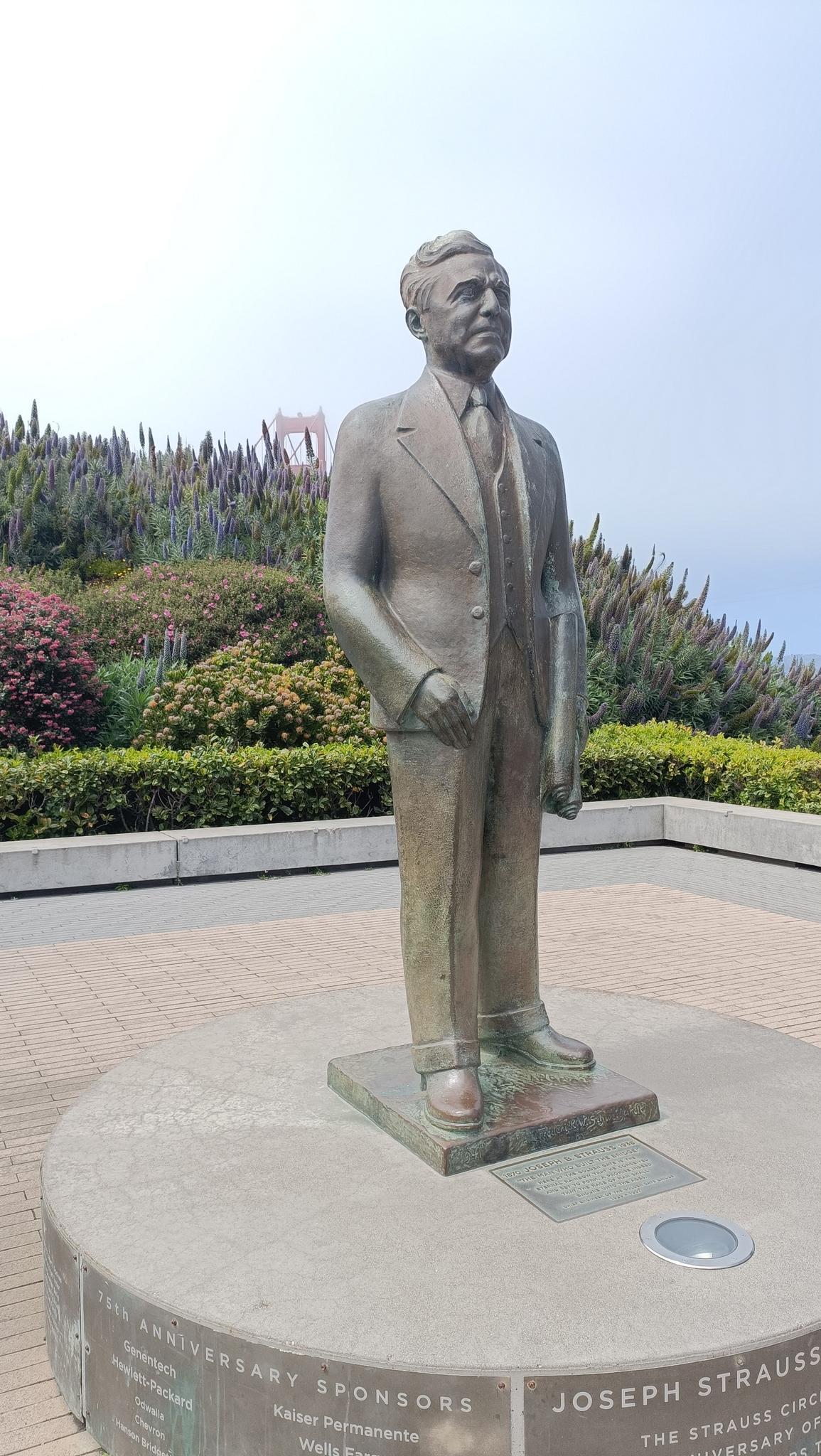 Bronze statue of a man in a suit, with green plants in background.
