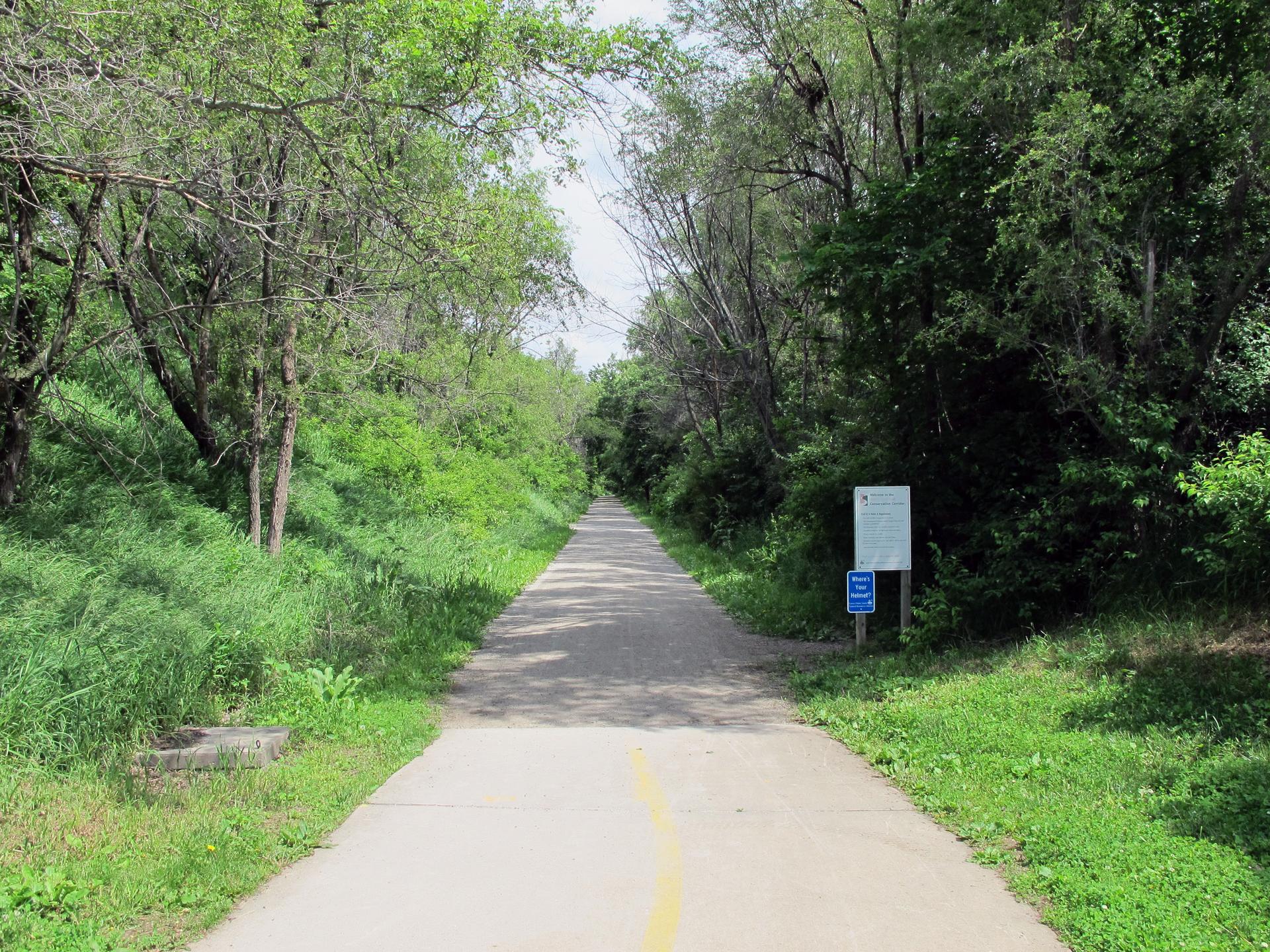 Paved path lined by dense green trees, with signs on the right, under bright skies.