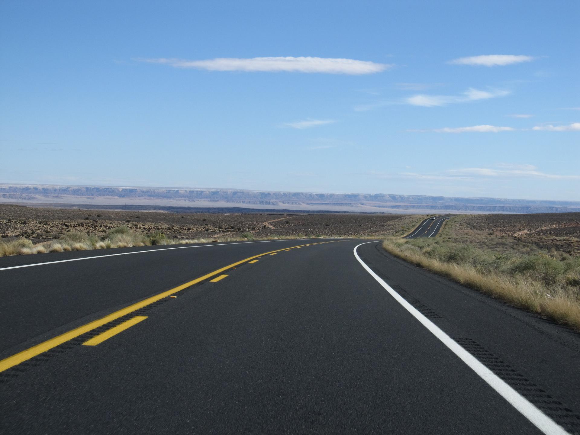 Empty two-lane road winding through a flat, arid landscape under a blue sky.