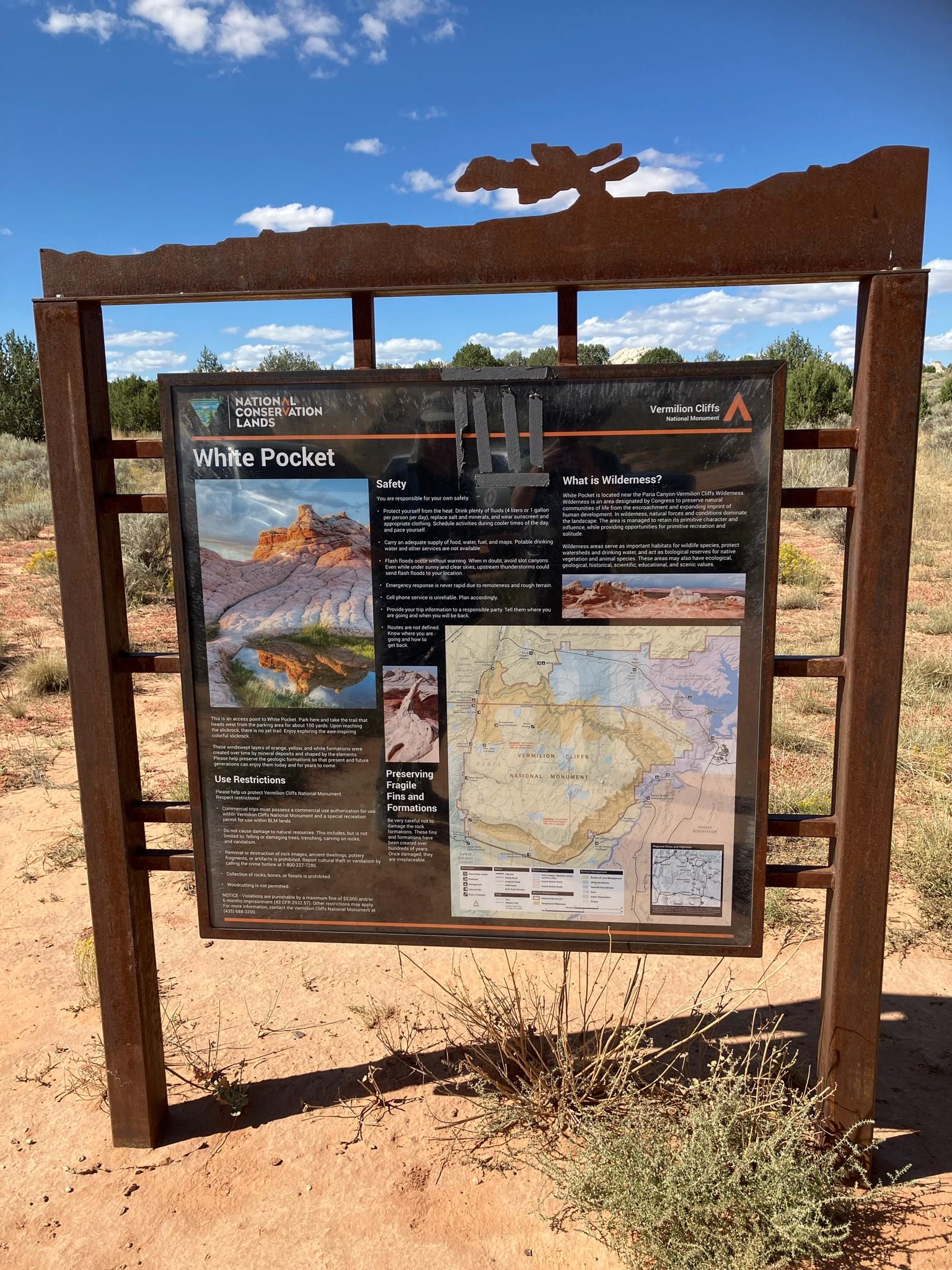 An informational sign with maps and text stands in a sandy, sparse desert. - White Pocket Trail