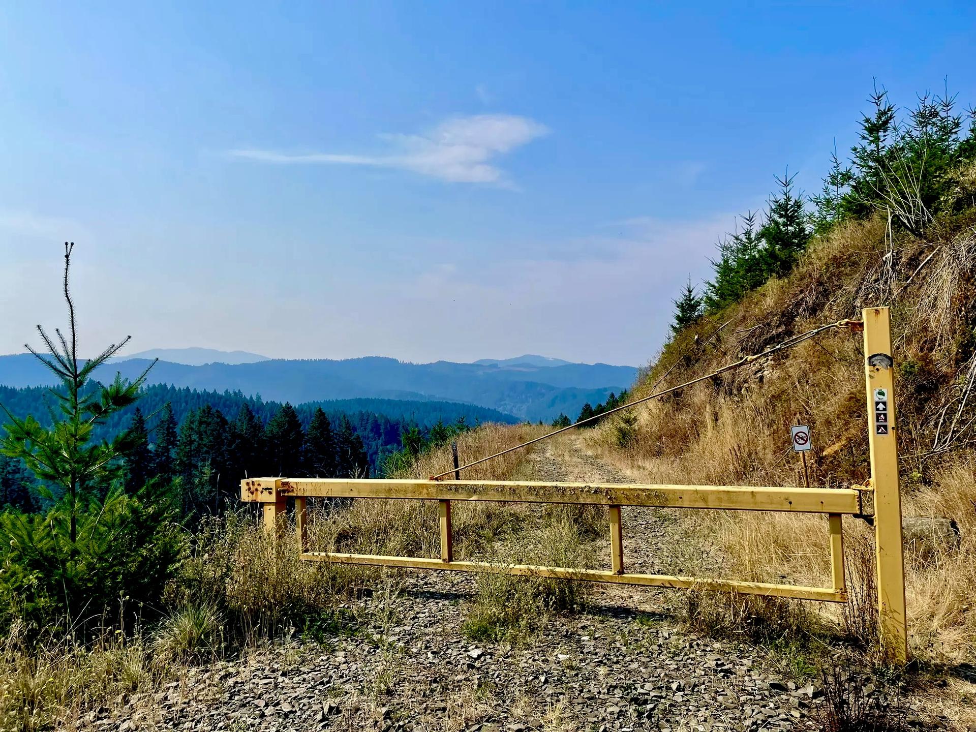 Yellow metal gate blocking a gravel road with forested hills beyond.