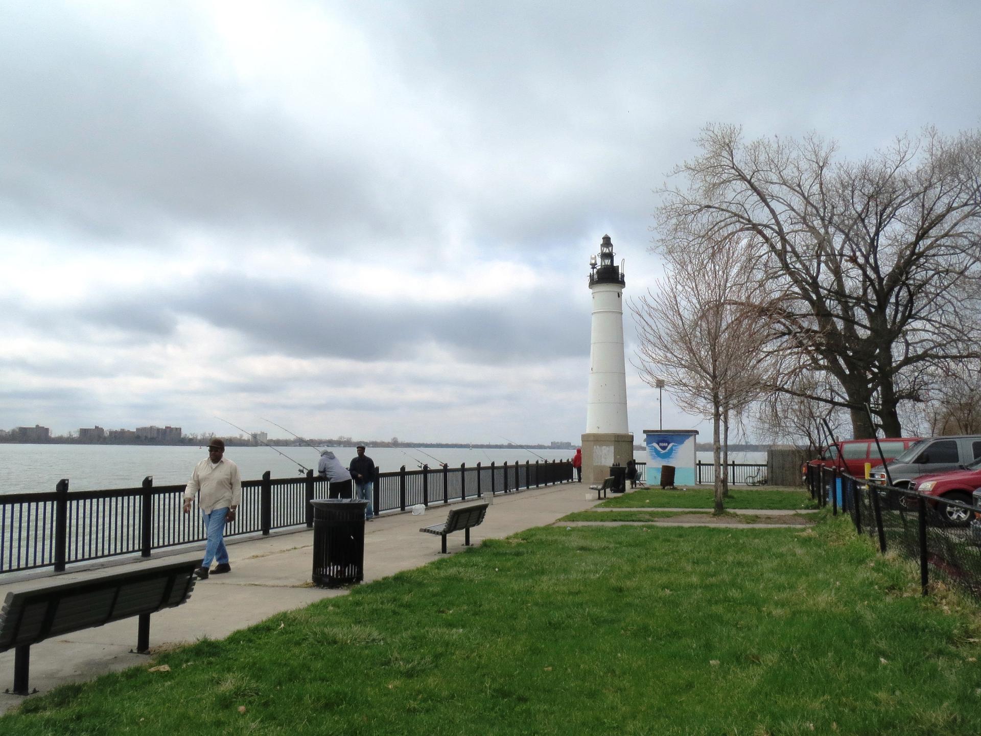 People walk and fish along a waterfront promenade with a white lighthouse under a cloudy sky.