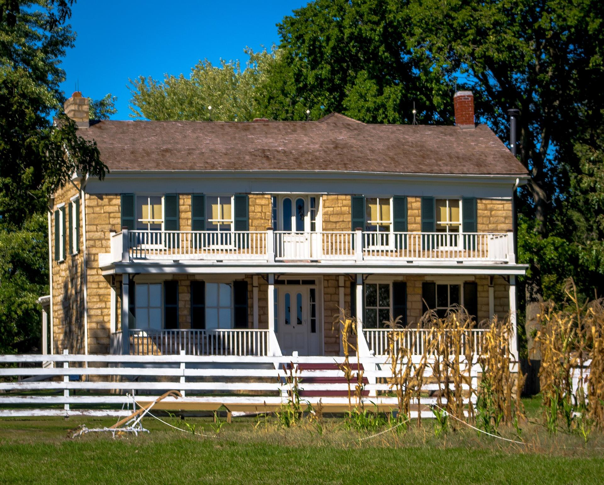 A historic stone house with a white porch and fence under a bright blue sky.