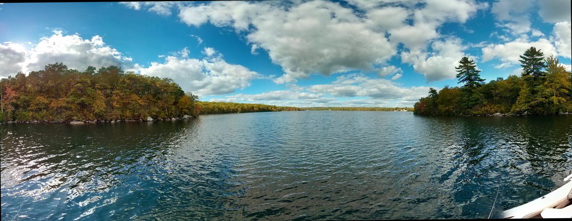 Panoramic view of a calm lake surrounded by autumn-colored forests under a blue sky.